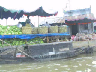 Khung cảnh tại một khu chợ nổi trên sông ở Cần Thơ, có vẻ là vào dịp cận Tết Nguyên Đán. Nhiều ghe, thuyền lớn nhỏ đang neo đậu hoặc di chuyển san sát nhau, tạo nên một không khí mua bán tấp nập. Trên các ghe thuyền chất đầy hàng hóa, đặc biệt là nông sản phục vụ Tết. Nổi bật là những chiếc ghe chở đầy dưa hấu được chất thành đống lớn, bên trên có các giỏ tre. Một số thuyền khác dường như đang chở các chậu hoa màu vàng, có thể là hoa cúc hoặc hoa mai. Con người đang thực hiện hoạt động mua bán, trao đổi hàng hóa ngay trên sông. Có thể thấy một người ngồi trên chiếc ghế nhựa màu đỏ đặt trên thuyền, bên cạnh là một động cơ máy dầu. Không có chữ viết nào có thể đọc được trong hình.