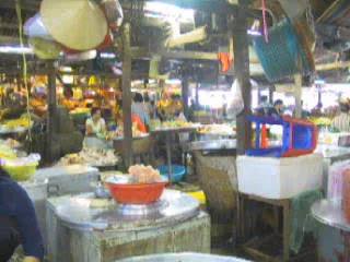 Hình ảnh ghi lại khung cảnh nhộn nhịp bên trong một khu chợ truyền thống, có thể là ở Cần Thơ. Không khí trong chợ rất đông đúc với nhiều người bán và người mua, cho thấy đây có thể là cảnh mua sắm chuẩn bị cho dịp Tết. Các tiểu thương, chủ yếu là phụ nữ, đang ngồi tại các quầy hàng của mình. Ở tiền cảnh, một người phụ nữ mặc áo màu xanh lam đang ngồi bán hàng. Trên quầy hàng trước mặt cô có một cái thau lớn màu đỏ đựng một loại thực phẩm màu hồng, có thể là thịt xay hoặc chả cá. Xung quanh là nhiều quầy hàng khác bày bán các loại thực phẩm đa dạng. Có thể thấy nhiều vật dụng quen thuộc của khu chợ được treo trên các cột và trần nhà, chẳng hạn như nón lá, chảo nấu ăn và rổ nhựa. Phía bên phải có một thùng xốp màu trắng. Không có biển hiệu hay chữ viết nào có thể đọc rõ trong ảnh.