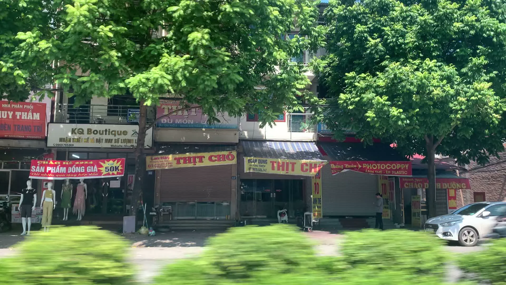 Cảnh quay ghi lại khung cảnh một con đường ở Việt Nam, được chụp từ một phương tiện đang di chuyển. Hai bên đường là một dãy các cửa hàng kinh doanh liền kề nhau dưới những tán cây xanh. Nhiều biển hiệu có thể được nhìn thấy rõ ràng, bao gồm: cửa hàng thời trang 'KQ BOUTIQUE' với thông tin 'NHẬN THIẾT KẾ & ĐẶT MAY SỐ LƯỢNG LỚN', nhà phân phối 'UY TRÂM', và một biểu ngữ 'CHÀO HÈ 2024 SẢN PHẨM ĐỒNG GIÁ 50K'. Đáng chú ý là có hai cửa hàng bán 'THỊT CHÓ' với các biển hiệu 'CẦY TƠ BẢY MÓN' và 'LẨU - NƯỚNG', một trong số đó đang đóng cửa cuốn. Ngoài ra còn có các cửa hàng dịch vụ khác như 'Photocopy', quán 'CHÁO DINH DƯỠNG', 'NHÀ THUỐC THƯ VÂN', và cửa hàng 'HOA TƯƠI PHƯƠNG THÀNH'. Hình ảnh cũng cho thấy một cột mốc bên đường ghi 'ĐOAN NỮ 27 KM'. Giao thông trên đường bao gồm ô tô và xe máy. Một số ma-nơ-canh đang trưng bày quần áo trước một cửa hàng.