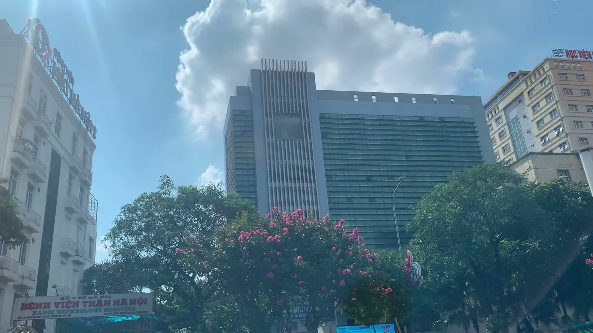 Đây là khung cảnh đường phố tại Hà Nội, được chụp từ một phương tiện đang di chuyển. Có nhiều tòa nhà cao tầng, hiện đại hai bên đường. Một biển hiệu lớn có thể được nhìn thấy rõ với các dòng chữ: 'SỞ Y TẾ HÀ NỘI', 'BỆNH VIỆN THẬN HÀ NỘI' (HANOI NEPHROLOGY HOSPITAL), và địa chỉ 'SỐ 70 NGUYỄN CHÍ THANH - ĐỐNG ĐA - HÀ NỘI'. Ở phía đối diện, một tòa nhà khác có chữ 'HỌC VIỆN' trên đỉnh. Dọc theo con đường là những hàng cây xanh và những cây hoa bằng lăng màu hồng. Một chiếc xe taxi màu trắng của hãng 'Taxi Group' cũng xuất hiện trong một bức ảnh. Bầu trời trong xanh, có nắng và mây trắng, cho thấy một ngày thời tiết đẹp. Hoạt động chính được ghi lại là di chuyển trên đường phố đô thị.