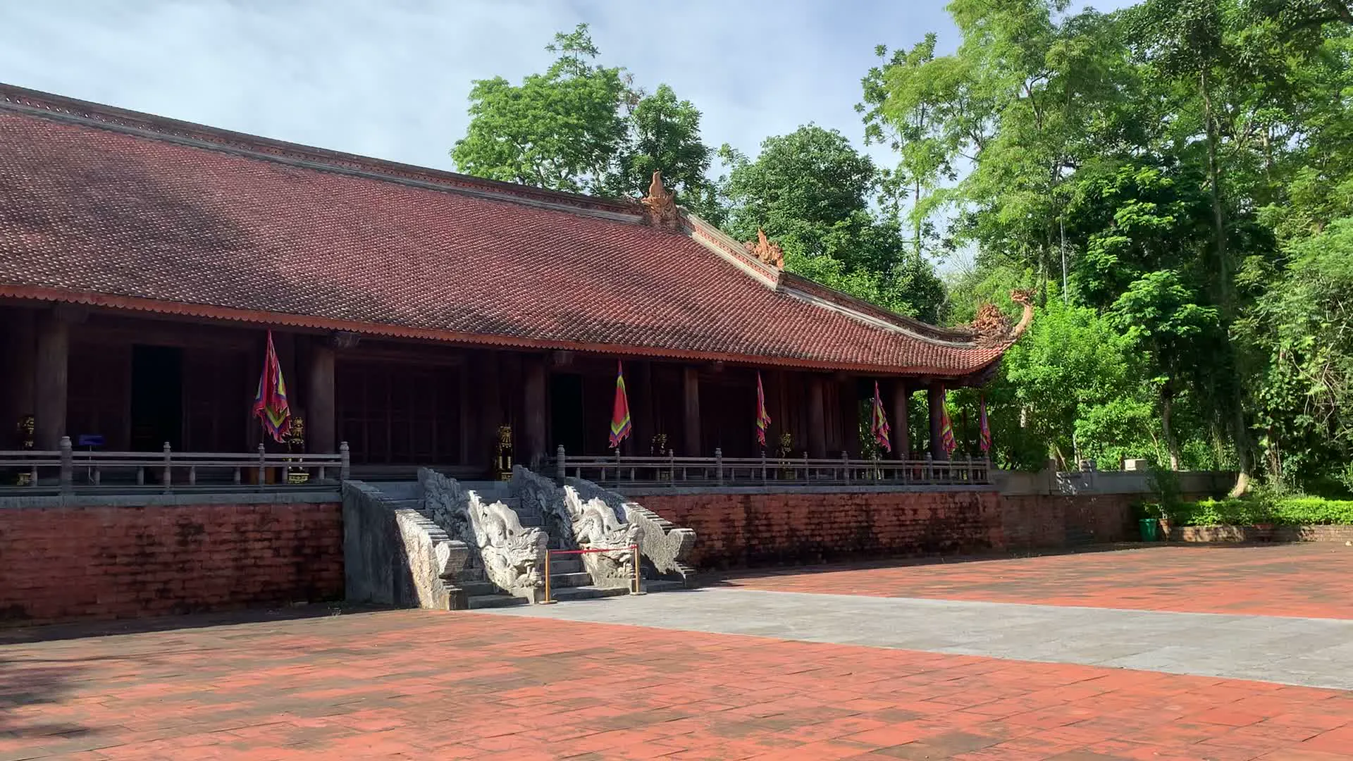 Bối cảnh là một khu di tích lịch sử rộng lớn, có khả năng là Khu di tích Lam Kinh, với kiến trúc cổ kính và không gian trang nghiêm. Hình ảnh cho thấy một ngôi điện thờ chính, có mái ngói đỏ và kết cấu bằng gỗ, được xây trên một nền cao. Lối lên điện có bậc thềm bằng đá với đôi rồng hoặc nghê đá được chạm khắc tinh xảo ở hai bên. Dọc theo hiên của ngôi điện có treo nhiều lá cờ hội ngũ sắc.。Phía trước điện là một khoảng sân rất rộng được lát hoàn toàn bằng gạch đỏ. Trong sân có một tấm biển nhỏ màu trắng nhưng không thể đọc rõ nội dung. Xung quanh khu vực là một không gian xanh mát với nhiều cây cối, nổi bật là một cây đa cổ thụ khổng lồ với bộ rễ phụ rủ xuống.。Hoạt động chính được ghi lại là việc tham quan của du khách. Một nhóm nhỏ gồm người lớn và trẻ em đang đi dạo trong sân, dưới bóng cây. Một người đàn ông khác đang đi một mình qua sân, hướng về phía ngôi điện. Không khí chung rất yên tĩnh và thanh bình, phù hợp với một địa điểm văn hóa, lịch sử.