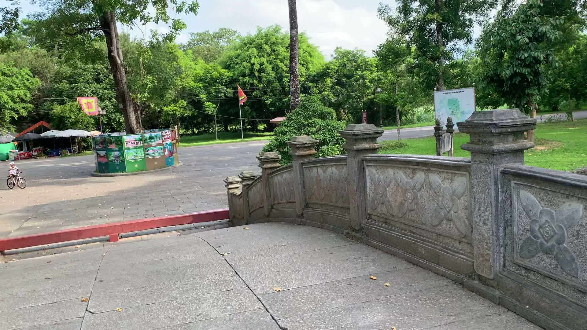 Bối cảnh là một khu di tích lịch sử với không gian rộng rãi, nhiều cây xanh và các lối đi được lát đá. Nổi bật trong các hình ảnh là một cây cầu bằng đá với thành cầu được chạm khắc hoa văn tinh xảo. Từ trên cầu nhìn xuống là một con đường lát đá dẫn vào một công trình kiến trúc cổ có mái ngói đỏ ở phía xa. Hai bên lối đi là hàng cây và bãi cỏ xanh mướt.。Trong khu vực có một người đàn ông lớn tuổi, có vẻ là du khách, đang đứng trên cầu. Ông đội mũ, mặc áo sơ mi kẻ, quần dài, đeo một chiếc túi màu đen và tay cầm máy ảnh. Ở phía xa, có các quán nhỏ có ô che, và cờ Tổ quốc cùng cờ lễ hội đang bay. Có một em bé đang đi xe đạp ở khoảng sân rộng. Ngoài ra, có một bảng thông tin lớn và một biển báo nhỏ màu nâu trên bãi cỏ, nhưng chữ viết trên đó không thể đọc được. Khung cảnh chung cho thấy đây là một hoạt động tham quan, vãn cảnh tại một địa điểm văn hóa, lịch sử.