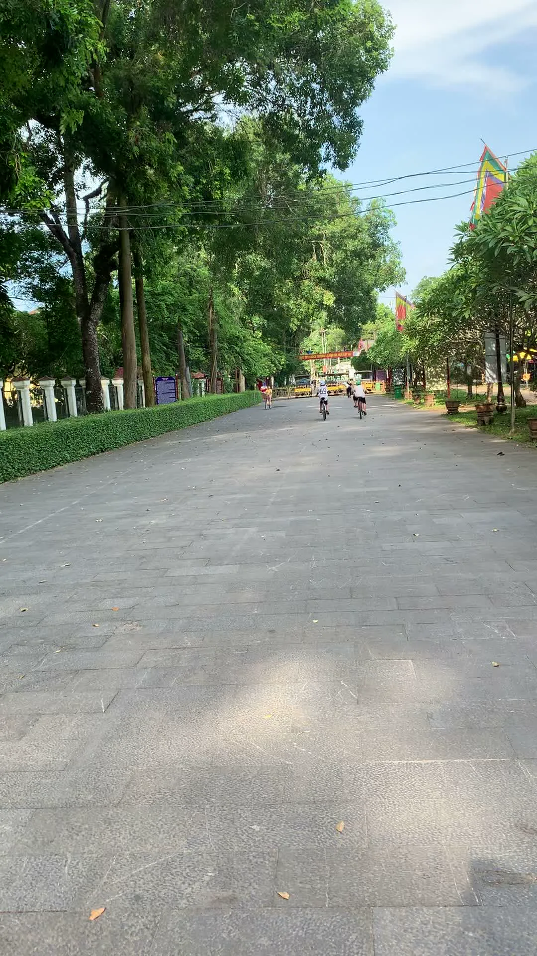 Cảnh quay ghi lại một con đường rộng lớn được lát bằng đá phiến màu xám, nằm trong một không gian thoáng đãng và xanh mát. Hai bên đường là những hàng cây cổ thụ cao lớn, phía bên trái có một hàng rào cây xanh được cắt tỉa gọn gàng và một hàng rào cột trắng. Một nhóm người, có vẻ là học sinh, đang đạp xe trên đường, di chuyển ra xa. Ở phía xa cuối con đường, có thể nhìn thấy một công trình kiến trúc giống như cổng vào. Phía bên phải có treo một lá cờ lễ hội màu đỏ. Có một biển báo màu xanh ở bên trái nhưng không thể đọc được chữ. Hoạt động chính là mọi người đang đạp xe và đi dạo để giải trí hoặc tập thể dục trong một khu vực công cộng, có thể là công viên hoặc khu di tích.