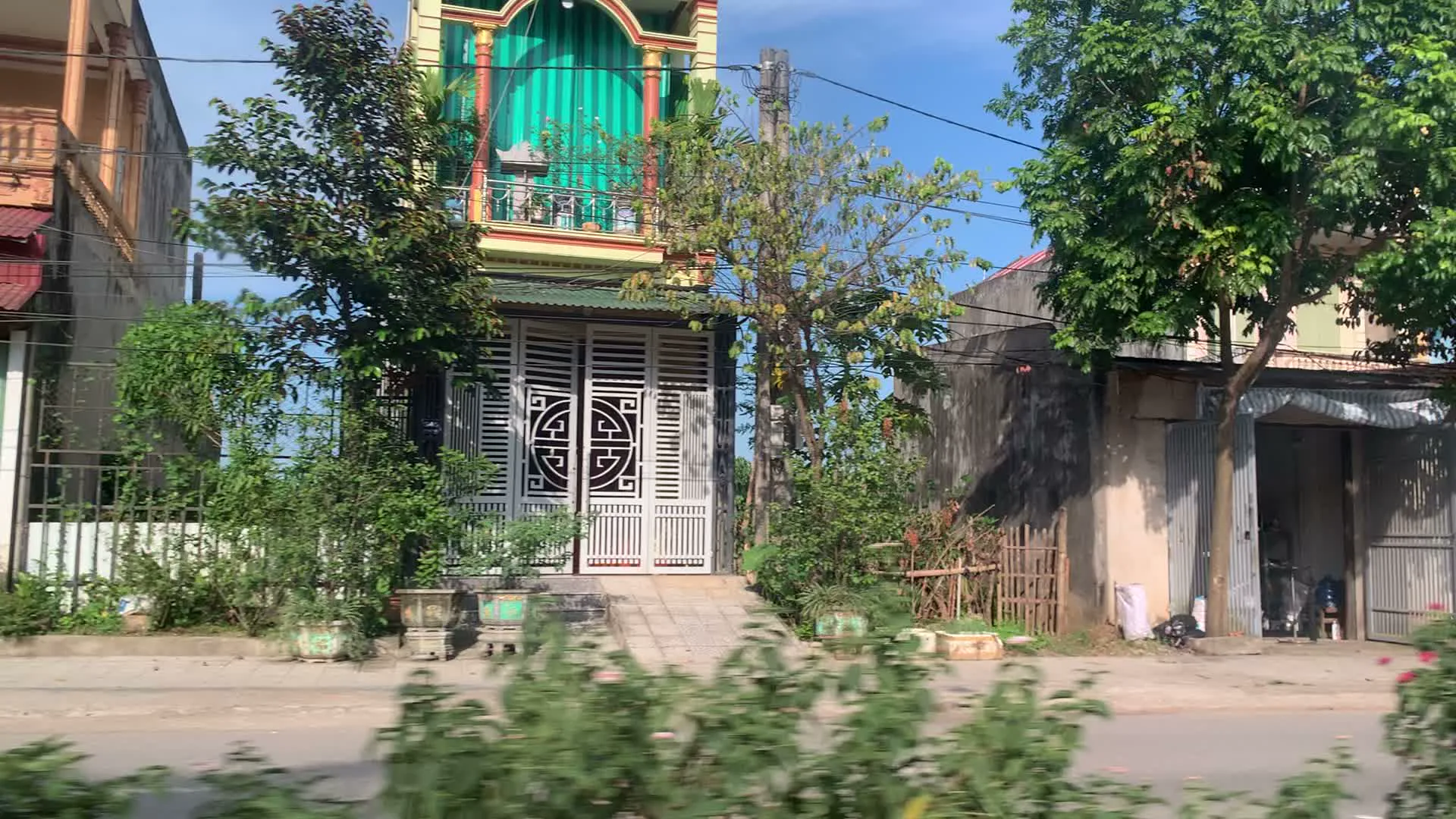 Quang cảnh dọc một con đường ở một khu dân cư tại Việt Nam, được chụp từ một phương tiện đang di chuyển. Các hình ảnh cho thấy những ngôi nhà ống nhiều tầng đặc trưng. Một ngôi nhà có mặt tiền màu xanh lá cây, ban công và cổng sắt màu trắng với hoa văn hình tròn. Cây cối và cây cảnh được trồng nhiều dọc theo con đường. Có một cổng chào lớn với họa tiết rồng, trên đó có dòng chữ "LAM SƠN" và "KÍNH CHÀO QUÝ KHÁCH". Bên cạnh cổng là một biểu ngữ dọc màu đỏ có các chữ "HOA BÌNH CÔNG LÝ ĐÁ GIAO DUYÊN". Một ngôi nhà khác màu xanh lam có cửa cuốn đang mở, để lộ một chiếc xe máy đang đỗ bên trong. Một lá cờ đỏ sao vàng của Việt Nam được treo dưới mái hiên sọc xanh. Trên cùng tuyến đường, có một cửa hàng với biển hiệu "121 VỊT QUAY HOÀNG KIM" và một tấm biển nhỏ hơn ghi "BIA HƠI". Khung cảnh tổng thể mô tả một khu vực sinh sống và kinh doanh nhỏ lẻ với đường nhựa, nhà cửa san sát và hệ thống dây điện chằng chịt trên cao dưới bầu trời xanh.