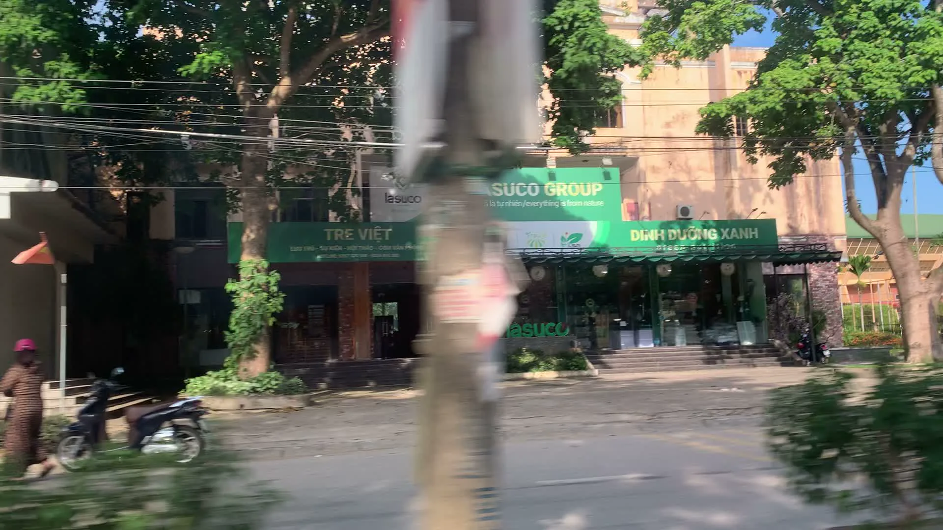 Đây là một đoạn video quay cảnh đường phố từ một phương tiện đang di chuyển. Khung cảnh cho thấy một con đường trải nhựa với cây xanh ở hai bên.。Trong phần đầu của video, có thể thấy một dãy nhà với các cửa hàng. Một cửa hàng nổi bật với biển hiệu lớn màu xanh lá cây ghi 'SUCO GROUP' và khẩu hiệu 'là tự nhiên / everything is from nature'. Ngay bên cạnh là một cửa hàng khác cũng có biển hiệu màu xanh lá cây ghi 'DINH DƯỠNG XANH'. Về phía bên trái là cửa hàng 'TRE VIỆT', trên biển hiệu có ghi 'ĐẶC SẢN - NÔNG SẢN - VẬT PHẨM...'. Phía trước các cửa hàng, có một người đang đi bộ trên vỉa hè và một chiếc xe máy đang đậu.。Sau đó, góc quay lia qua một khu vực khác, cho thấy một khuôn viên rộng lớn được rào lại bằng hàng rào bê tông sơn màu đỏ và nâu, có cổng sắt. Bên trong hàng rào là một tòa nhà lớn nhiều tầng với mái màu xanh lá. Xung quanh tòa nhà và dọc theo hàng rào có nhiều cây cao, bao gồm cả cây cau. Nhiều cột điện với hệ thống dây điện chằng chịt cũng xuất hiện trong cảnh quay.