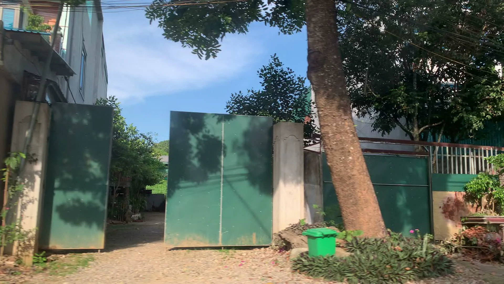 Các hình ảnh ghi lại khung cảnh dọc một con đường ở khu vực dường như là nông thôn hoặc ngoại ô, được chụp từ một phương tiện đang di chuyển. Bức ảnh đầu tiên cho thấy một lối vào một khu nhà với cổng sắt lớn màu xanh lá cây đang mở. Phía sau cổng là một ngôi nhà tầng và nhiều cây cối. Phía trước, bên phải cổng là một thân cây to và một thùng rác nhỏ màu xanh. Con đường là đường đất sỏi. Trong các hình ảnh tiếp theo, một chiếc ô tô màu xanh lam lướt qua. Bên đường có một ngôi nhà cấp 4 lợp mái tôn, phía trước có treo lá cờ Tổ quốc màu đỏ. Ở phía xa có thể thấy một cột điện cao thế. Một cảnh khác cho thấy một tòa nhà có cửa cuốn lớn bằng kim loại màu xanh lá cây. Bên cạnh tòa nhà có một số biển hiệu quảng cáo, trong đó có một biển màu xanh dương và một biển màu vàng, nhưng chữ viết trên đó bị mờ và không thể đọc được do xe đang di chuyển. Cảnh vật xung quanh có nhiều cây xanh và cỏ dại mọc ven đường.