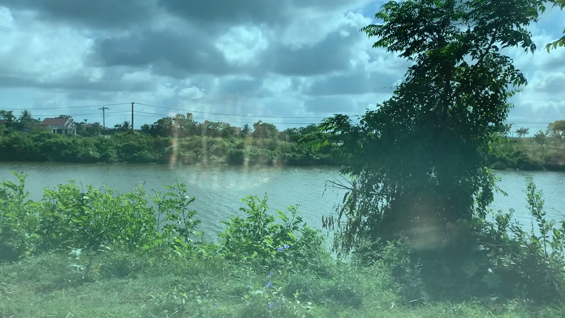 Đây là khung cảnh một vùng nông thôn, có một con sông rộng nước màu xanh đục chảy qua. Hai bên bờ sông được bao phủ bởi cây cối và bụi rậm xanh tốt. Phía xa bên kia sông là những lùm cây rậm rạp, thấp thoáng có một ngôi nhà mái ngói đỏ. Bầu trời nhiều mây, tạo nên một khung cảnh yên bình. Có các cột điện và đường dây điện chạy dọc theo bờ sông ở phía xa. Hình ảnh có vẻ được chụp từ bên trong một chiếc xe đang di chuyển, thể hiện qua sự thay đổi góc nhìn và các vệt sáng phản chiếu trên kính.