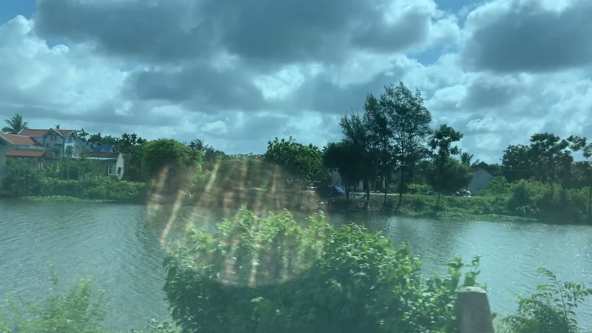 Bức ảnh ghi lại cảnh quan vùng nông thôn ven sông, được chụp từ một phương tiện đang di chuyển. Ở phía trước là một con sông hoặc ao lớn. Dọc hai bên bờ là cây cối và bụi rậm xanh tươi. Phía xa, bên kia bờ sông, có thể thấy những ngôi nhà dân với mái ngói nhiều màu sắc nằm xen kẽ giữa các lùm cây. Bầu trời có mây. Hình ảnh có một chút phản chiếu ánh sáng, cho thấy nó có thể được chụp qua cửa kính.