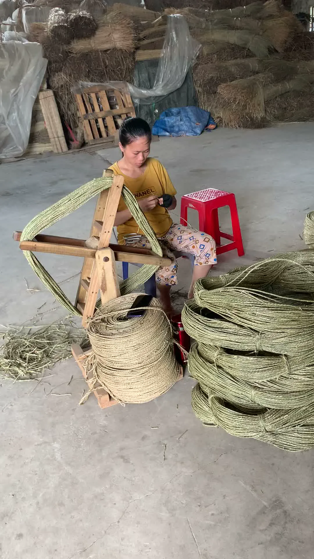 Trên áo phông màu vàng của người phụ nữ có in chữ "HANEL". Bối cảnh là một nhà xưởng hoặc nhà kho thủ công với sàn bê tông. Một người phụ nữ đang ngồi trên chiếc ghế nhựa màu đỏ, thực hiện công việc cuốn sợi cói. Cô đang sử dụng một khung quay bằng gỗ để quấn các sợi cói khô thành những cuộn dây lớn và đều đặn. Bên cạnh cô là nhiều cuộn sợi cói đã hoàn thành được xếp chồng lên nhau. Dưới sàn và ở phía sau là nguyên liệu cói khô, với những kiện cói lớn được xếp chồng lên cao. Đây là một công đoạn trong quy trình sản xuất các sản phẩm thủ công từ cây cói, một nghề truyền thống đặc trưng của vùng Nga Sơn.