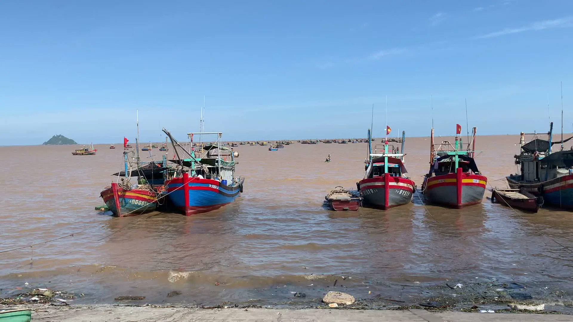 Bối cảnh là một cảng cá ven biển sầm uất, có khả năng là ở xã Ngư Lộc, tỉnh Thanh Hóa. Hình ảnh cho thấy rất nhiều tàu cá, ghe thuyền đang neo đậu trên vùng nước biển có màu nâu đặc trưng của phù sa. Bầu trời quang đãng, trong xanh. Các tàu cá được sơn màu sắc nổi bật như xanh và đỏ, trên nhiều tàu có cắm cờ Tổ quốc. Có thể đọc được số hiệu đăng ký trên một số tàu, chẳng hạn như "TH 91819 TS", trong đó "TH" là ký hiệu của tỉnh Thanh Hóa. Phía xa trên biển có một hòn đảo nhỏ. Cảnh tượng này phản ánh hoạt động nghề cá sôi động. Các tàu thuyền đang neo đậu, có thể là đang nghỉ ngơi, chờ ra khơi hoặc vừa cập bến sau chuyến đánh bắt. Bờ kè được làm bằng bê tông và sỏi đá lẫn vỏ sò. Một hình ảnh khác ghi lại cảnh sinh hoạt và buôn bán trên bờ. Một chiếc xe tải nhỏ màu trắng có thùng đông lạnh đang đỗ ngay trước một cửa hàng. Hai người phụ nữ đội nón lá đang làm việc, có thể là đang giao hoặc nhận hàng hóa (nhiều khả năng là hải sản) từ xe tải vào cửa hàng. Gần đó có một chiếc sọt tre lớn và một chiếc mẹt tre. Cảnh này cho thấy hoạt động thương mại, phân phối sản phẩm sau đánh bắt, một phần không thể thiếu trong đời sống của người dân làng chài.