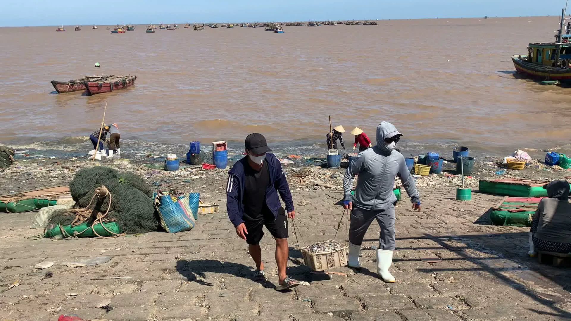 Đây là khung cảnh tại một bến cá hoặc cảng cá ven biển tấp nập, có khả năng là ở khu vực Ngư Lộc. Bờ kè được xây bằng bê tông, dốc xuống mép nước, là nơi người dân tập trung để mua bán và sơ chế hải sản. Nước biển có màu nâu đục. Trên biển, rất nhiều tàu thuyền đánh cá (tàu, ghe) đang neo đậu san sát nhau. Một số tàu thuyền có sơn màu sặc sỡ và ghi số hiệu, có thể đọc được một phần như "TH 91...", là ký hiệu của tỉnh Thanh Hóa.。Hoạt động chính diễn ra là cảnh lao động của các ngư dân. Có những người đàn ông đang hợp sức khiêng các giỏ đầy ắp hải sản vừa đánh bắt được đi lên từ mép nước. Nhiều phụ nữ đội nón lá đang ngồi hoặc đứng ở mép nước để phân loại cá, tôm từ các giỏ lớn vào những chiếc xô, chậu nhựa. Khung cảnh rất nhộn nhịp, đặc trưng của một cảng cá khi tàu thuyền vừa cập bến.。Các vật thể xuất hiện nhiều trong ảnh bao gồm: tàu thuyền đánh cá, thuyền thúng, nón lá, các loại giỏ, xô, chậu nhựa màu xanh dương và xanh lá để đựng hải sản, và các đống lưới đánh cá. Dọc bờ kè có khá nhiều rác thải. Ở phía xa ngoài khơi có thể nhìn thấy một hòn đảo nhỏ.