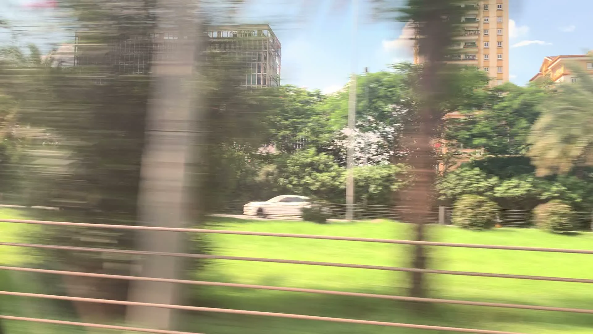 Cảnh quan được ghi lại từ một phương tiện đang di chuyển, do đó hình ảnh bị mờ vì chuyển động. Ở phía trước là lan can đường bằng kim loại. Nhìn qua lan can là một không gian xanh với bãi cỏ và nhiều loại cây cối, bao gồm cả cây cọ. Một chiếc ô tô màu trắng có thể được nhìn thấy trên một con đường song song. Ở phía sau là các tòa nhà cao tầng, dường như là các khu chung cư hoặc văn phòng trong một khu đô thị. Có một biển báo màu xanh ở phía xa nhưng không thể đọc được nội dung. Toàn bộ khung cảnh cho thấy đây là một con đường hiện đại, có thể là đường cao tốc hoặc đại lộ.