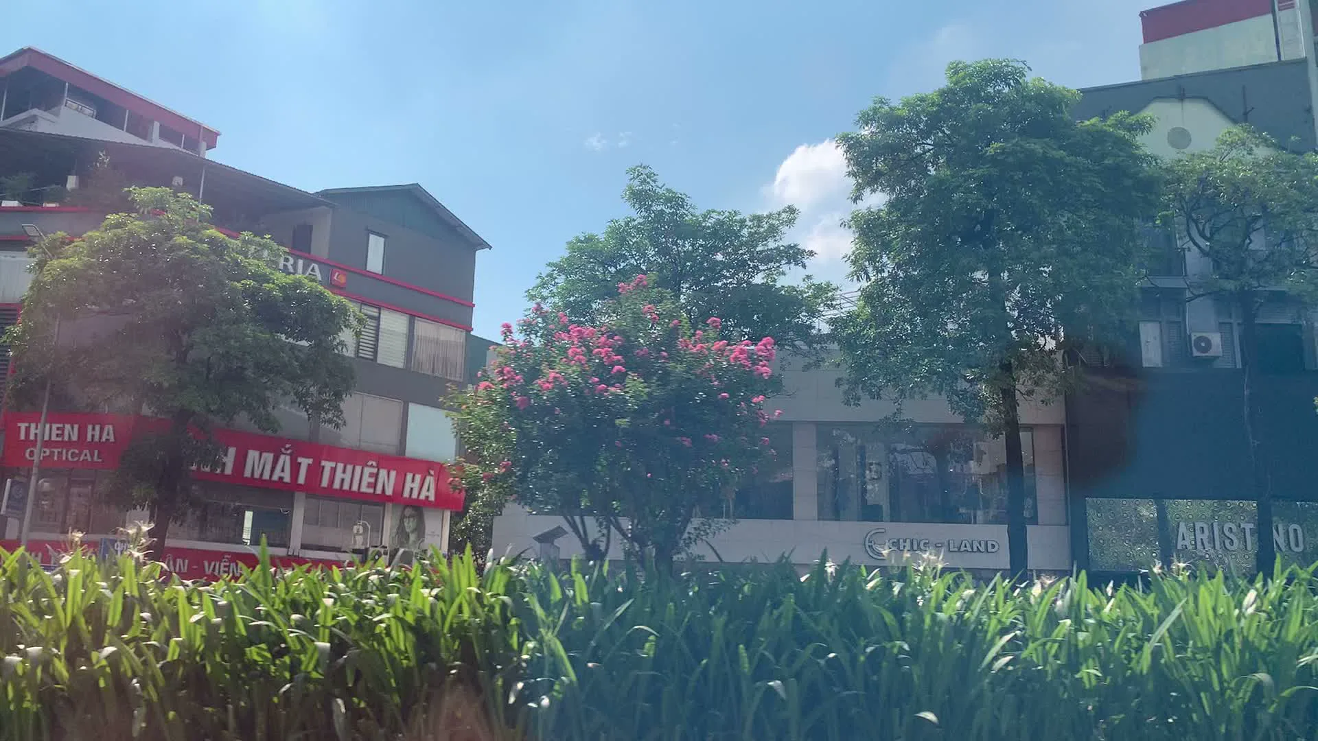 Bối cảnh là một con đường phố đô thị, được quay từ một phương tiện đang di chuyển. Bầu trời trong xanh, có vài gợn mây. Hai bên đường là những tòa nhà cao tầng với nhiều cửa hàng và cây xanh. Có thể nhận diện được các biển hiệu của một số cửa hàng như "KÍNH MẮT THIÊN HÀ", "THIEN HA OPTICAL", "CHIC-LAND", và "ARISTINO". Cảnh vật bao gồm nhiều cây xanh, trong đó có một cây bằng lăng đang nở hoa màu hồng tím. Một người đang đi xe máy trên đường, đội mũ bảo hiểm và đeo khẩu trang. Khung cảnh cho thấy hoạt động giao thông và thương mại thường ngày trên một con phố ở Việt Nam.