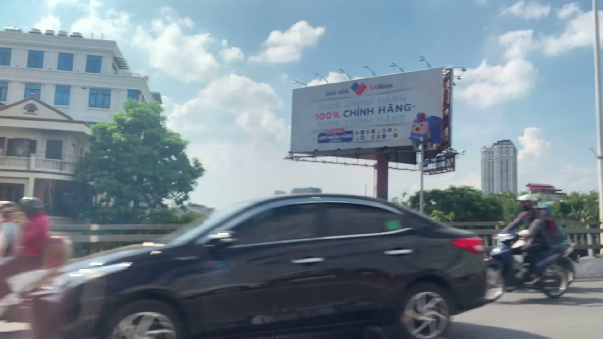 Hình ảnh ghi lại cảnh giao thông trên một cây cầu hoặc cầu vượt trong một khu đô thị. Có một biển quảng cáo lớn của 'VnShop' với nội dung 'MUA SẮM' và '100% CHÍNH HÃNG'. Nhiều phương tiện đang lưu thông trên đường, bao gồm một chiếc ô tô màu đen, một người đi xe máy và hai người khác đang đi trên một chiếc xe tay ga màu cam. Phía sau là các tòa nhà, cây cối và một tòa nhà cao tầng ở phía xa. Lan can cầu được sơn sọc đỏ và trắng. Bầu trời trong xanh và có mây.