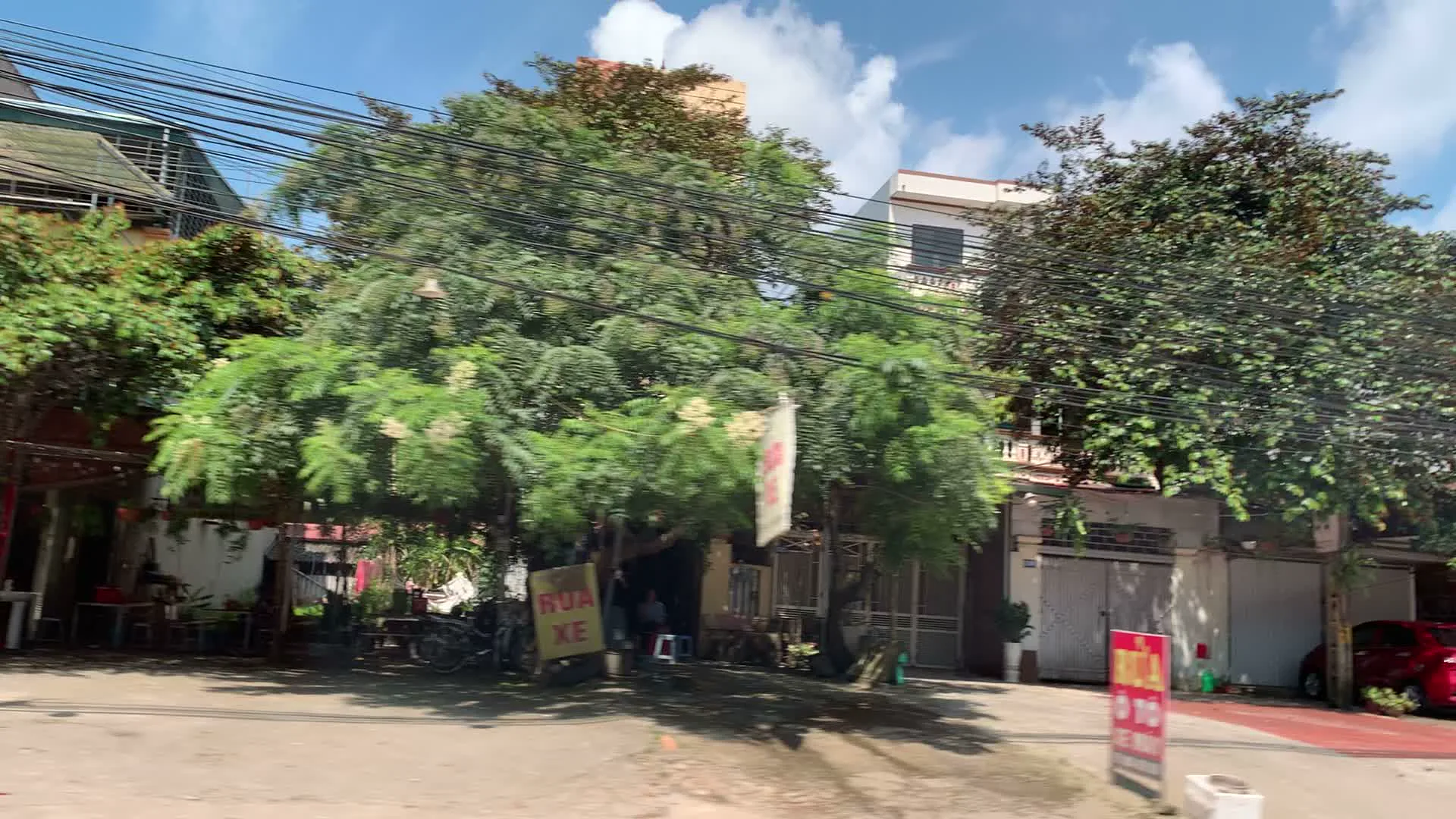 Cảnh quay từ một phương tiện đang di chuyển trên một con đường ở khu vực đô thị, có thể là ở Sơn Tây. Hai bên đường là những ngôi nhà ống nhiều tầng san sát nhau và rất nhiều cây xanh. Bầu trời trong xanh, có mây. Có thể thấy rõ các biển hiệu của một cửa hàng ven đường. Biển hiệu màu đỏ với chữ trắng ghi 'RỬA XE', 'THAY DẦU', 'BƠM LỐP', cho thấy đây là một tiệm rửa và sửa chữa xe máy. Một vài chiếc xe máy đang đỗ gần đó. Bên lề đường còn có một đống bao tải màu trắng. Khung cảnh thể hiện một góc phố yên tĩnh với các hoạt động kinh doanh nhỏ lẻ thường ngày ở Việt Nam.