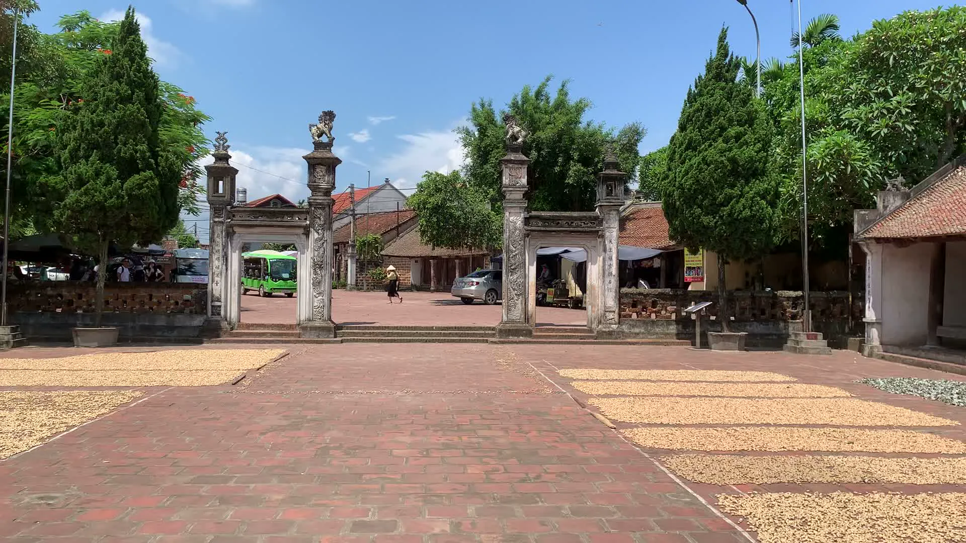 Bối cảnh là một không gian kiến trúc truyền thống của Việt Nam, có khả năng là một ngôi đình làng hoặc di tích lịch sử tại khu vực Sơn Tây. Hình ảnh bên ngoài cho thấy một khoảng sân rộng lát gạch đỏ, nơi người dân đang phơi nông sản (trông giống như lạc hoặc đỗ) trên các tấm bạt. Lối vào chính là một cổng tam quan cổ được xây dựng công phu bằng đá, trên đỉnh các trụ cổng có đặt tượng nghê. Xuyên qua cổng có thể thấy một phần của ngôi làng với nhà cửa, cây xanh, một chiếc xe ô tô và một chiếc xe điện du lịch màu xanh lá. Một người phụ nữ đội nón lá đang đi trong sân. Bên trong công trình là một gian thờ chính. Toàn bộ kết cấu được làm bằng gỗ với các cột gỗ lớn. Bàn thờ được bày biện trang trọng với các đồ thờ cúng như bình hoa và mâm quả. Nổi bật ở hai bên là một đôi hạc thờ bằng đồng mạ vàng, mỗi con đứng trên lưng rùa, được đặt trong các bình gốm sứ lớn. Sàn nhà được lát gạch và có một tấm chiếu cói trải ở phía trước. Có một hòm công đức đặt trên một chiếc ghế nhỏ. Chữ viết được ghi nhận: Trên hòm công đức có dòng chữ 'HÒM CÔNG ĐỨC'.