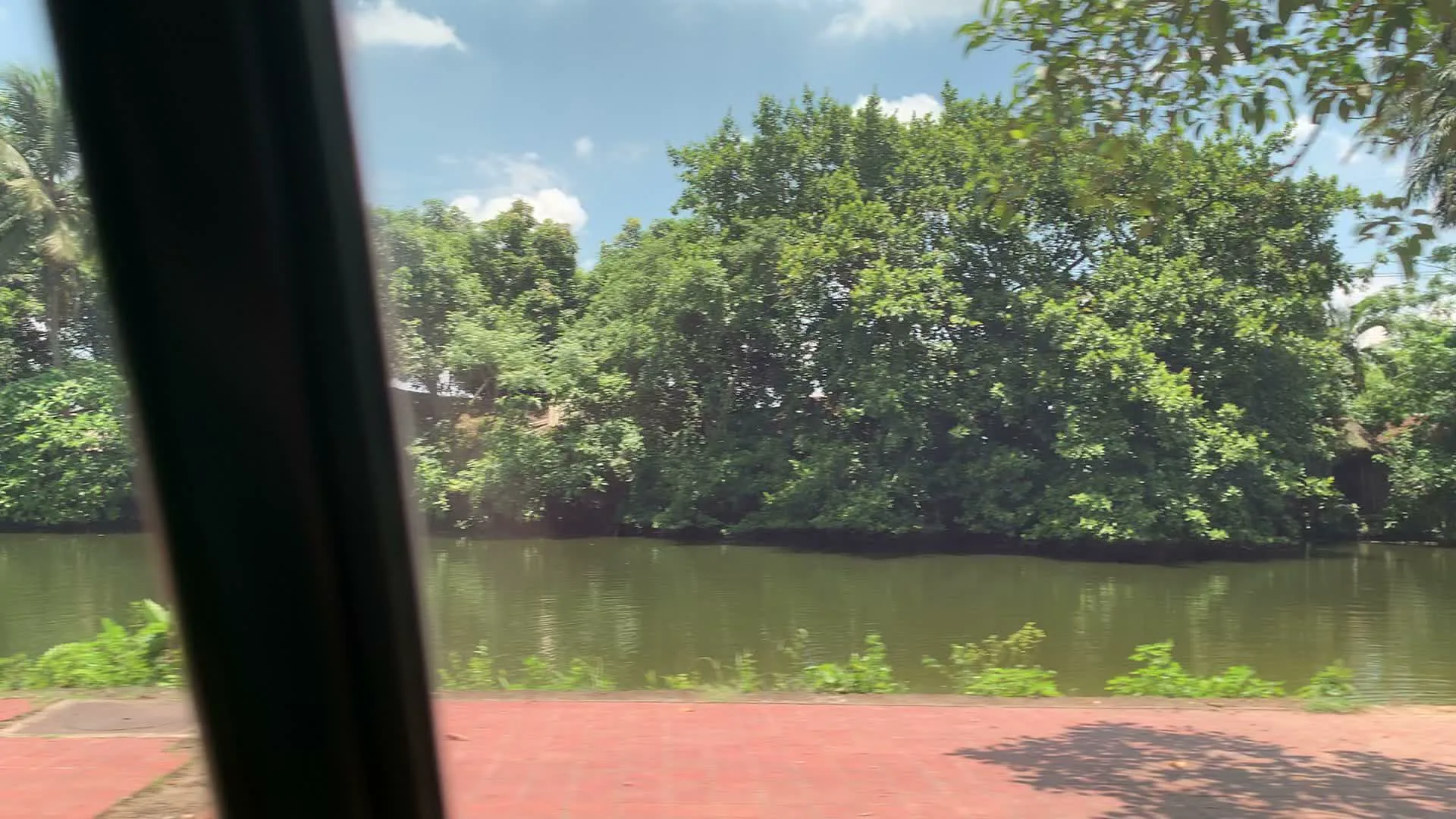 Cảnh quay được ghi lại từ một phương tiện đang di chuyển dọc theo một con đường ven ao hoặc hào nước. Phong cảnh đặc trưng với nhiều cây cối xanh tươi mọc um tùm quanh bờ ao. Dưới lòng ao, một người phụ nữ đội nón lá đang đứng ở mép nước, có vẻ như đang mò ốc hoặc bắt cá, bên cạnh là hai chiếc xô đặt trên bờ. Ven đường có một quán nước nhỏ dưới một chiếc ô lớn màu đen, trên tủ kính có biển hiệu ghi "NƯỚC MÍA SIÊU SẠCH". Gần đó, có một vài chiếc xe đạp đang đỗ, trong đó có một chiếc gắn biển "THUÊ XE ĐẠP ĐÔI", cho thấy đây có thể là một khu vực giải trí hoặc du lịch.