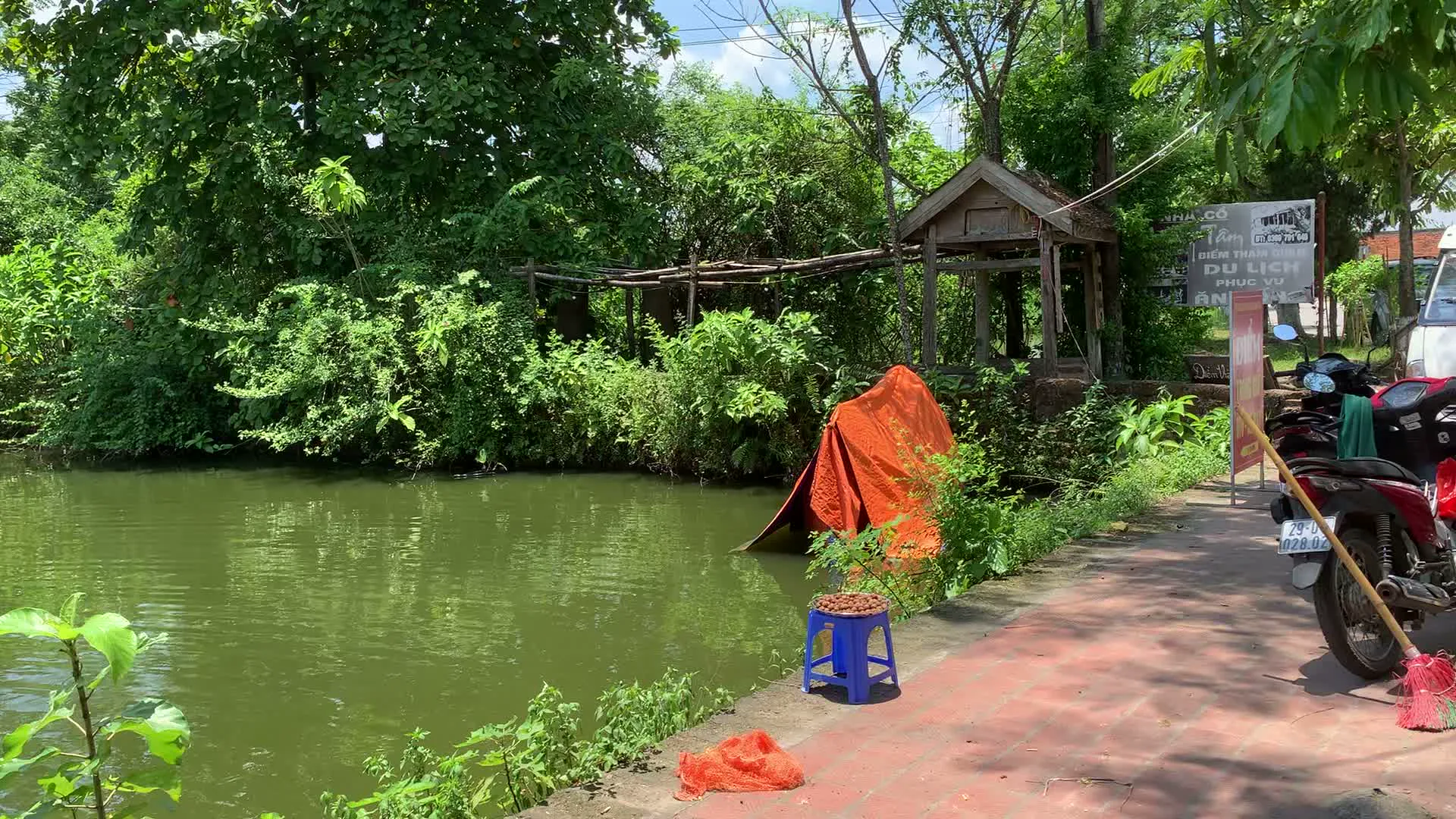 Các hình ảnh mô tả khung cảnh tại một ngôi làng ở vùng nông thôn, có khả năng là làng cổ Đường Lâm ở Sơn Tây, dựa trên các biển hiệu. Bối cảnh chung bao gồm một con ao hoặc hào nước làng với cây cối xanh tươi, bao quanh bởi một con đường lát gạch đỏ.。Hình ảnh đầu tiên chụp một góc yên tĩnh bên bờ ao. Có một biển hiệu với nội dung 'NHÀ CỔ', 'ĐIỂM THAM QUAN', 'DI TÍCH LỊCH SỬ PHÚC VŨ' và logo 'BIA HÀ NỘI'. Gần đó, có một chiếc ghế đẩu nhựa màu xanh và một ít nông sản (trông giống hạt dẻ) được bày bán. Một chiếc xe máy màu đỏ đang đậu trên vỉa hè.。Hình ảnh thứ hai cho thấy một người đang cúi xuống bên bờ ao, dùng rổ nhựa màu đỏ để rửa hoặc đãi thứ gì đó trong nước. Một chiếc xe đạp được dựng trên con đường lát gạch gần đó. Cảnh này thể hiện một hoạt động sinh hoạt thường ngày của người dân địa phương.。Hình ảnh thứ ba cho thấy một khu vực đông đúc hơn với một quán hàng nhỏ ven đường được che bởi hai chiếc ô lớn. Một nhóm người, có vẻ là khách du lịch, đang tụ tập quanh quán. Ở phía sau, có thể thấy cổng và biển hiệu của 'TRẠM Y TẾ XÃ ĐƯỜNG LÂM', xác nhận địa điểm là xã Đường Lâm.