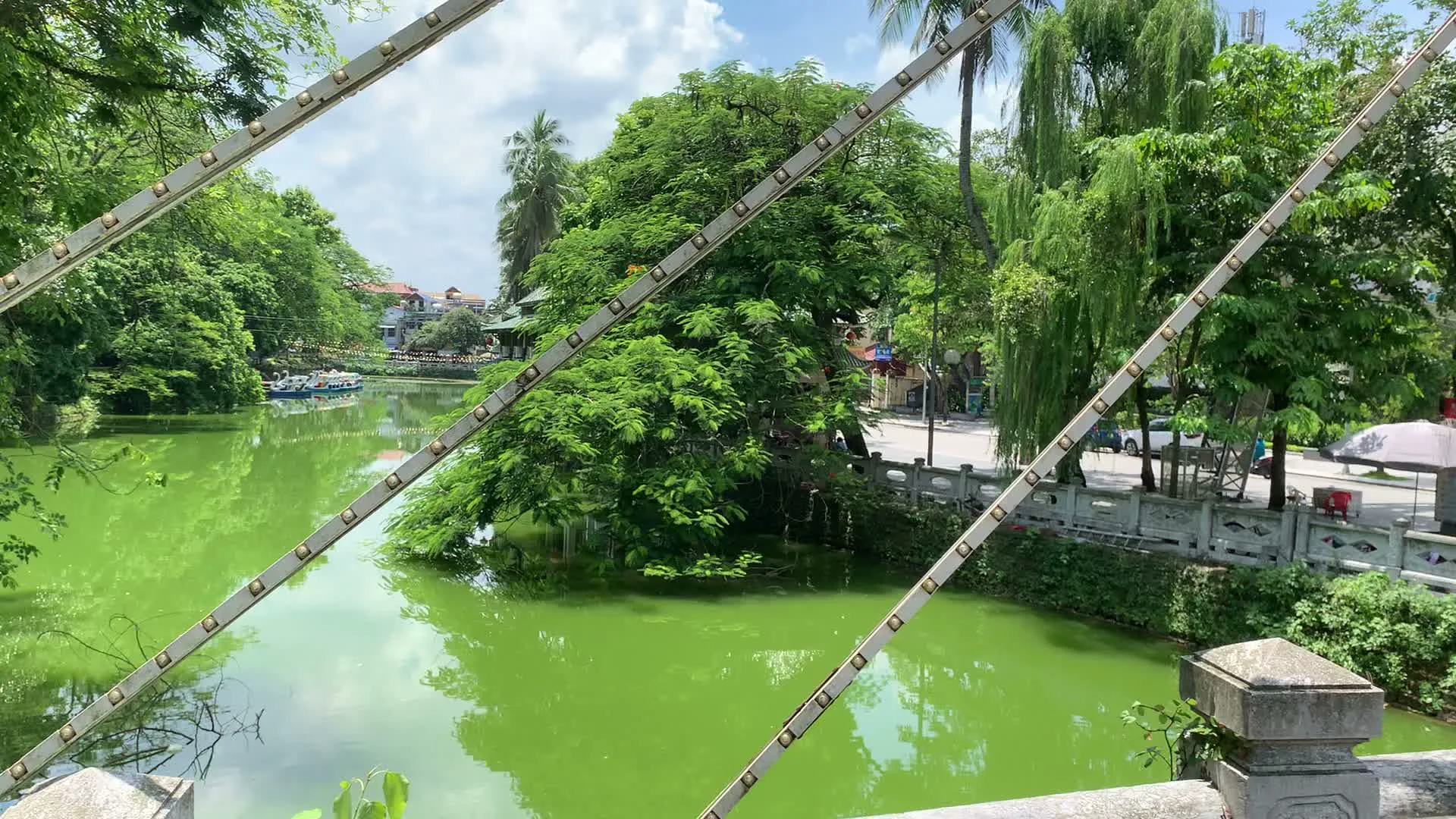 Đây là khung cảnh tại một hồ nước hoặc hào nước trong một khu vực có nhiều cây xanh, có thể là công viên hoặc khu di tích. Các bức ảnh được chụp từ trên một cây cầu có kết cấu độc đáo với các dây cáp kim loại chéo và lan can bằng đá.。Mặt nước hồ có màu xanh lục đặc trưng, phản chiếu bóng cây và bầu trời. Xung quanh hồ là rất nhiều cây cối xanh tươi, bao gồm cả cây liễu và cây dừa. Ven hồ có một con đường đi bộ.。Trong các bức ảnh, có thể thấy một nhóm người, một số mặc áo đỏ, đang đi dạo trên con đường ven hồ. Ở phía xa, có các tòa nhà mang nét kiến trúc truyền thống và một chiếc thuyền du lịch màu xanh đang đậu trên mặt hồ. Hoạt động chính là tham quan, ngắm cảnh của người dân và du khách. Không có chữ viết nào có thể đọc được.