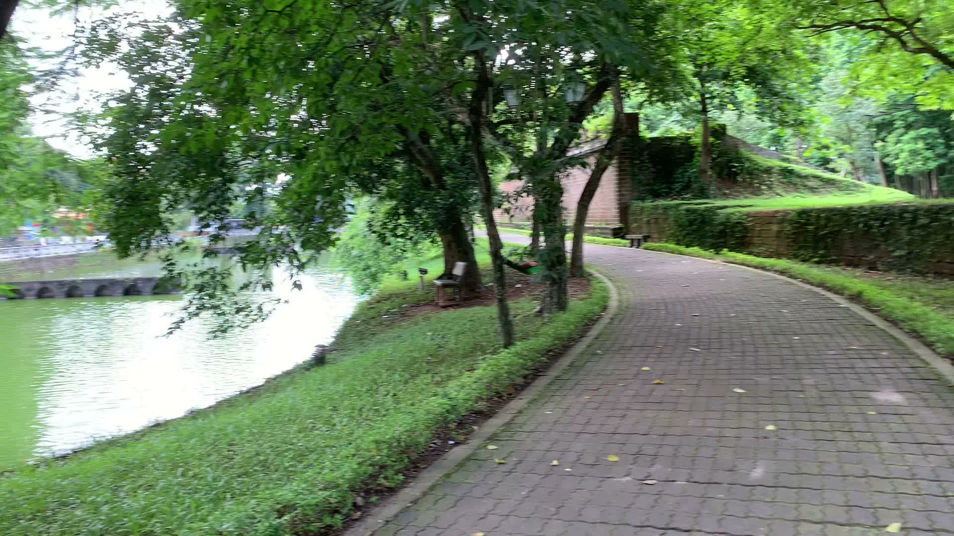 Khung cảnh tại một khu di tích lịch sử, có thể là một thành cổ, với một con đường đi bộ lát gạch uốn lượn. Con đường này chạy dọc theo một con hào hoặc hồ nước có màu xanh lục. Hai bên đường và ven bờ hào là những bãi cỏ xanh và rất nhiều cây cổ thụ cành lá xum xuê. Dưới bóng cây có đặt những chiếc ghế dài để khách nghỉ chân, và có một người đang nằm thư giãn trên chiếc võng mắc giữa hai cây. Ở phía xa, bắc qua hào nước là một cây cầu đá cổ có nhiều nhịp hình vòm. Phía bên kia con đường là một bức tường thành cổ, rêu phong. Có một tấm biển nhỏ gắn trên thân cây nhưng không thể đọc được chữ.