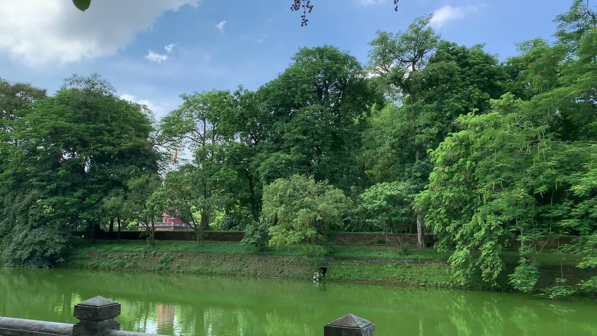 Đây là khung cảnh một hồ nước và con đường liền kề tại một khu vực đô thị. Hình ảnh cho thấy một hồ nước có màu xanh lục, được bao quanh bởi bờ kè và rất nhiều cây xanh tươi tốt. Dọc theo bờ hồ là một con đường với vỉa hè lát gạch, có lan can bằng đá. Trên vỉa hè có một số xe máy và một chiếc xe đạp đang đỗ. Một vài cây xanh được trang trí bằng đèn lồng nhiều màu sắc. Phía xa, bên kia đường là các tòa nhà và cửa hàng, nhưng các biển hiệu không thể đọc rõ. Bầu trời trong xanh, có mây trắng. Khung cảnh chung yên bình, thể hiện một góc của cuộc sống đô thị hòa quyện với thiên nhiên.