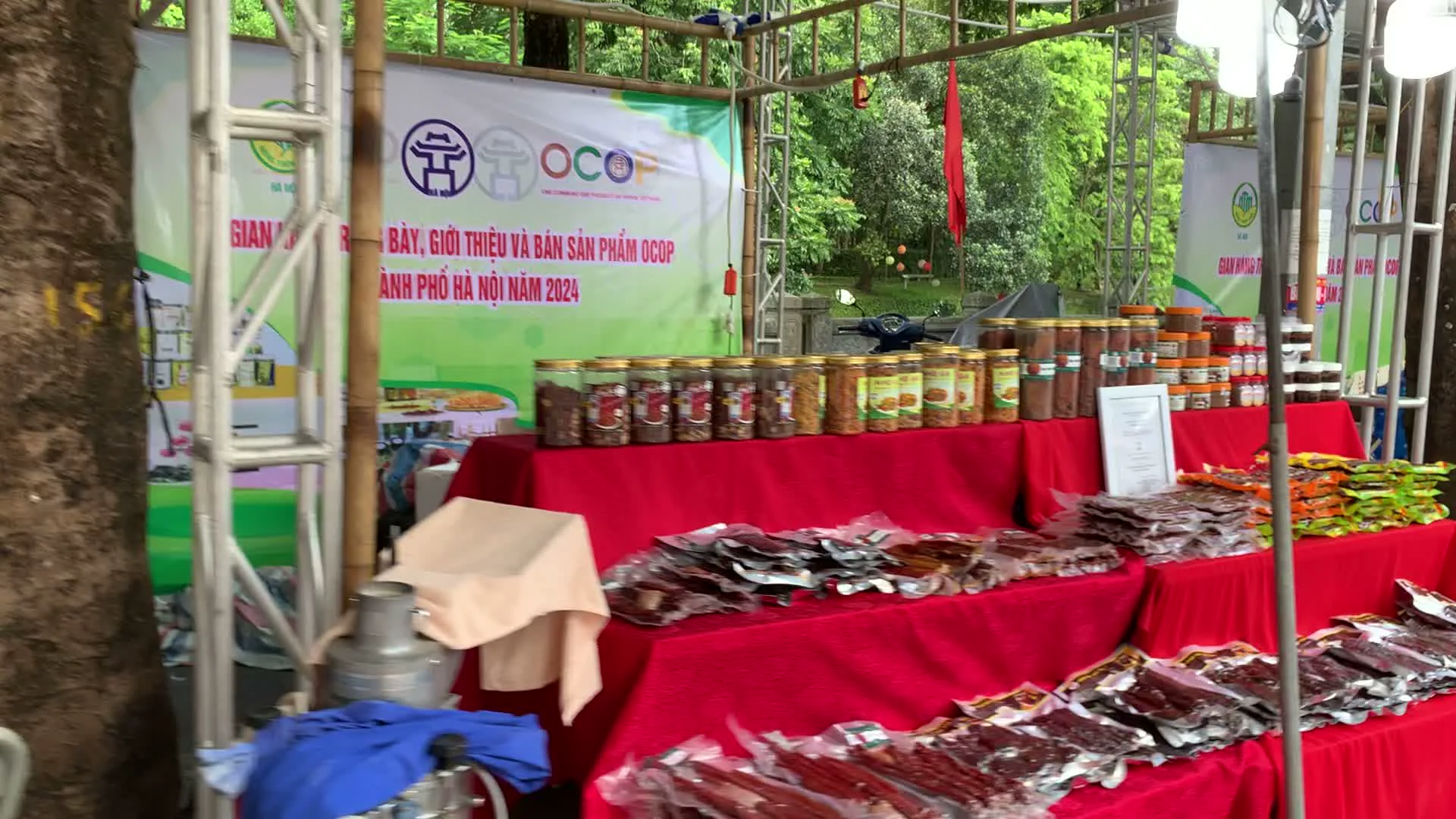 Bối cảnh là một hội chợ ngoài trời với nhiều gian hàng được dựng lên dưới các khung sắt và mái bạt. Xung quanh có nhiều cây xanh lớn, cho thấy địa điểm có thể là một công viên hoặc không gian công cộng. Các gian hàng đang trưng bày và bán nhiều loại sản phẩm khác nhau, chủ yếu là thực phẩm và đặc sản.。Chữ viết trên các tấm phông bạt:。- Một tấm phông lớn có nội dung: "GIAN HÀNG TRƯNG BÀY, GIỚI THIỆU VÀ BÁN SẢN PHẨM OCOP THÀNH PHỐ HÀ NỘI NĂM 2024", cùng với logo OCOP (Mỗi xã một sản phẩm) và logo của Hà Nội.。- Một tấm biển hiệu màu đỏ phía trước một quầy hàng ghi: "DOANH NGHIỆP TƯ NHÂN GIÒ CHẢ ƯỚC LỄ", "Trụ sở chính: Ước Lễ - Thanh Oai - Hà Nội" và số điện thoại.。- Các bao bì sản phẩm có chữ "BLACK BAG CHIA".。Hoạt động và vật thể:。- Các gian hàng được bày trên bàn phủ khăn màu đỏ và xanh dương.。- Sản phẩm được trưng bày bao gồm: các loại thịt khô đóng gói hút chân không, các loại hạt và thực phẩm khô đựng trong hũ thủy tinh, giò chả, hạt chia, các loại gia vị đóng chai, và các hộp thạch hoặc sữa chua.。- Có một chiếc cân điện tử màu xanh và một thớt gỗ trên bàn của quầy bán giò chả.。- Một tủ kính trưng bày các sản phẩm trông giống như vòng tay thủ công.。- Có một vài người bán hàng và khách tham quan. Một người phụ nữ mặc áo hồng đang đứng uống nước. Một người đàn ông đang ngồi nghỉ phía sau quầy hàng.