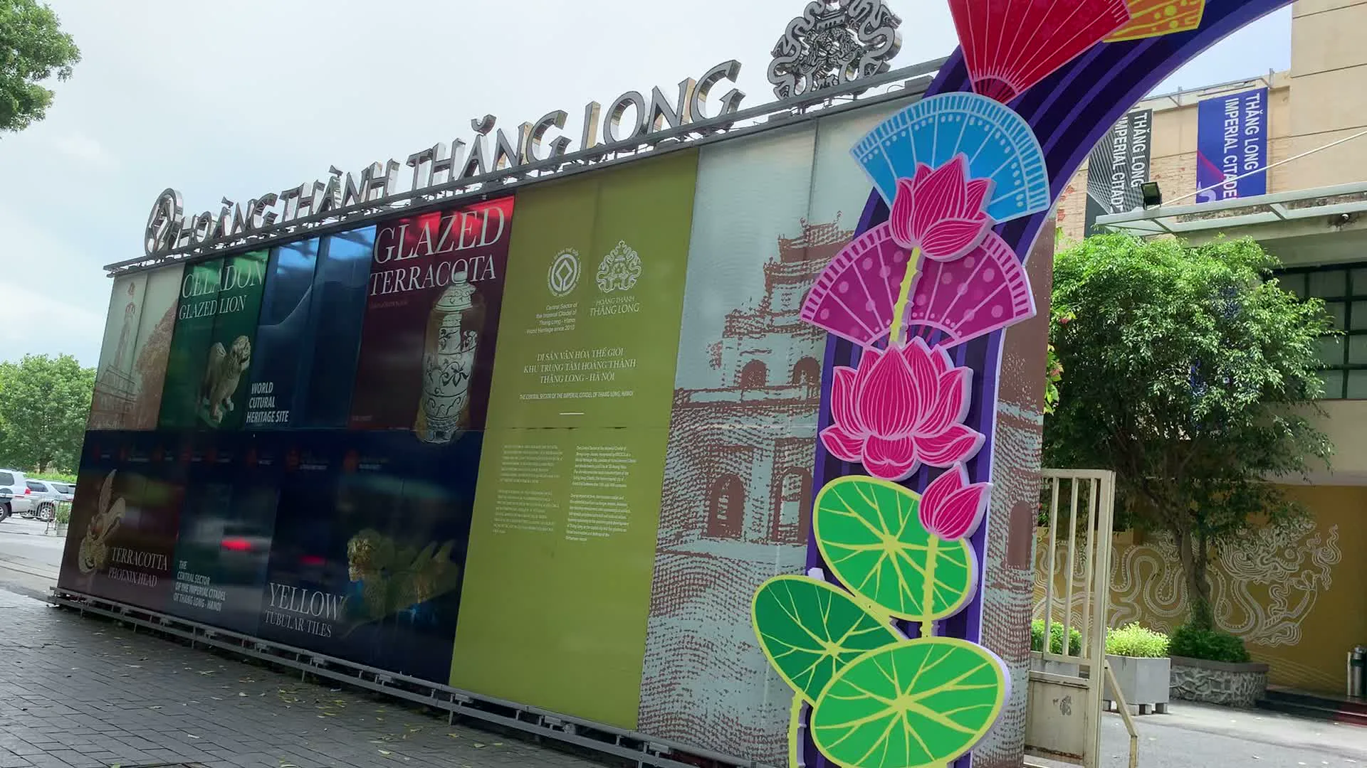 Đây là khu vực lối vào của Hoàng thành Thăng Long ở Hà Nội. Có một cổng chào lớn được trang trí công phu với các họa tiết hoa sen và quạt giấy nhiều màu sắc. Phía trên cổng có dòng chữ kim loại lớn "HOÀNG THÀNH THĂNG LONG". Lối vào khu vực bên trong có biển hiệu rõ ràng chỉ dẫn "NƠI BÁN VÉ / TIẾP KHÁCH THAM QUAN / TICKET OFFICE". Xung quanh được bố trí nhiều tấm pa-nô, áp phích quảng cáo cho các sự kiện và triển lãm. Một tấm áp phích lớn quảng bá cho sự kiện "Tết Đoan Ngọ / Thăng Long xưa / Đoan Ngọ Festival in Thang Long". Một tấm khác giới thiệu cuộc trưng bày "TRƯNG BÀY BÁU VẬT HOÀNG CUNG THĂNG LONG". Các pa-nô khác giới thiệu các hiện vật di sản bằng tiếng Anh như "GLAZED LION", "GLAZED TERRACOTA", "WORLD CULTURAL HERITAGE SITE". Các biển hiệu cũng xác nhận đây là "DI SẢN VĂN HÓA THẾ GIỚI KHU TRUNG TÂM HOÀNG THÀNH THĂNG LONG - HÀ NỘI", do "TRUNG TÂM BẢO TỒN DI SẢN THĂNG LONG - HÀ NỘI" quản lý. Trong ảnh có một người đàn ông đang ngồi trên xe máy và một vài du khách khác đứng gần đó, cho thấy đây là không gian công cộng ở lối vào một địa điểm du lịch văn hóa.