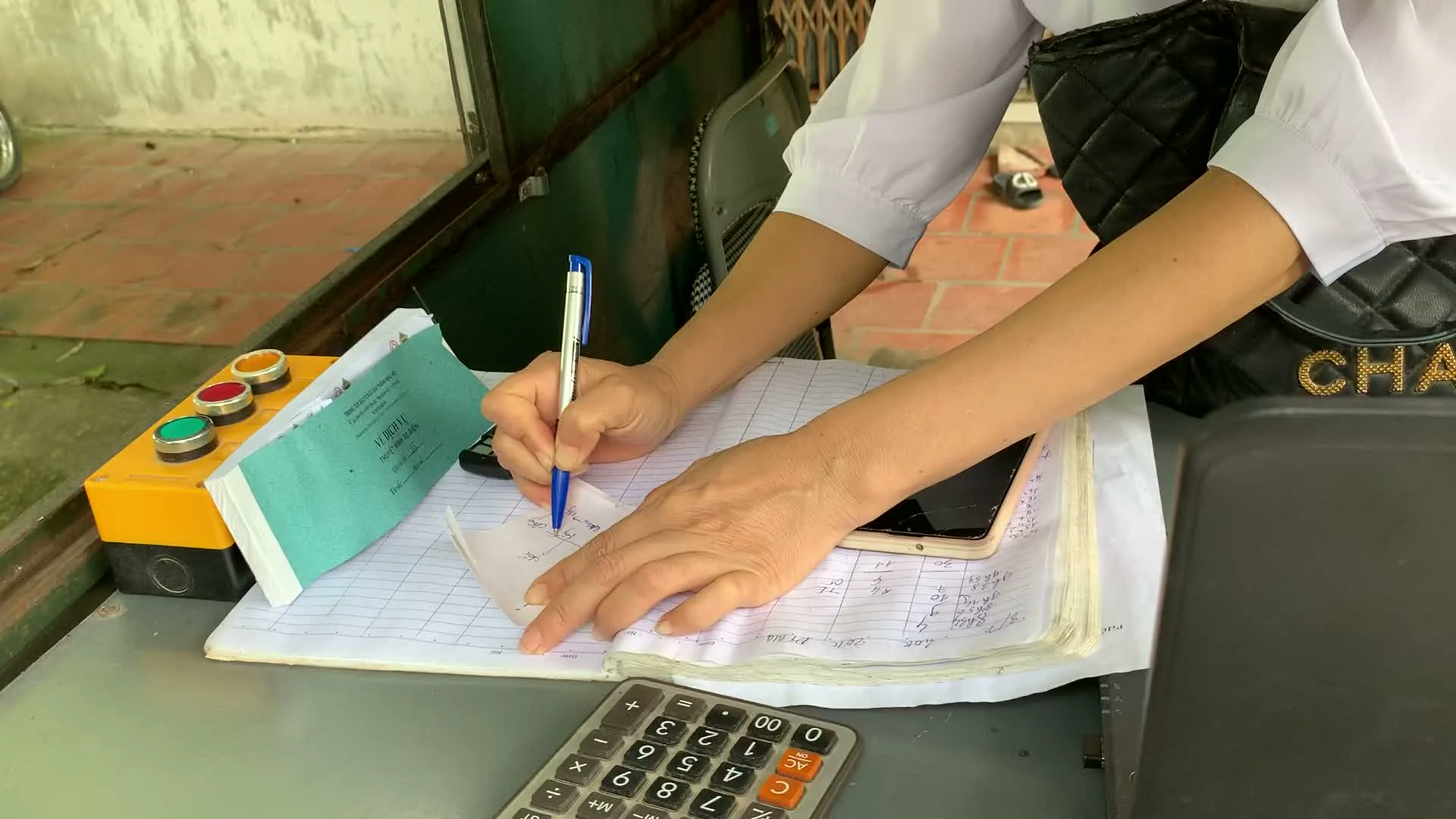 Một người phụ nữ mặc áo sơ mi trắng đang làm việc tại một quầy giấy. Cô ấy đang dùng bút bi để ghi chép các con số lên một mảnh giấy nhỏ, có vẻ như là một hóa đơn hoặc phiếu ghi nhận. Trên bàn làm việc có một cuốn sổ kế toán lớn với nhiều số liệu viết tay, một chiếc máy tính cầm tay, một chiếc điện thoại thông minh và một chiếc túi xách màu đen. Đặc biệt, có một cuốn sổ nhỏ màu xanh lá cây với tiêu đề có thể đọc được là "GIẤY CHỨNG NHẬN KIỂM DỊCH VẬN CHUYỂN ĐỘNG VẬT, SẢN PHẨM ĐỘNG VẬT TRONG NƯỚC". Gần đó là một hộp điều khiển công nghiệp màu vàng với các nút bấm màu đỏ, vàng và xanh lá. Bối cảnh cho thấy đây có thể là một trạm cân hoặc trạm kiểm dịch, nơi người phụ nữ đang thực hiện công việc ghi sổ sách, tính toán và xử lý giấy tờ.