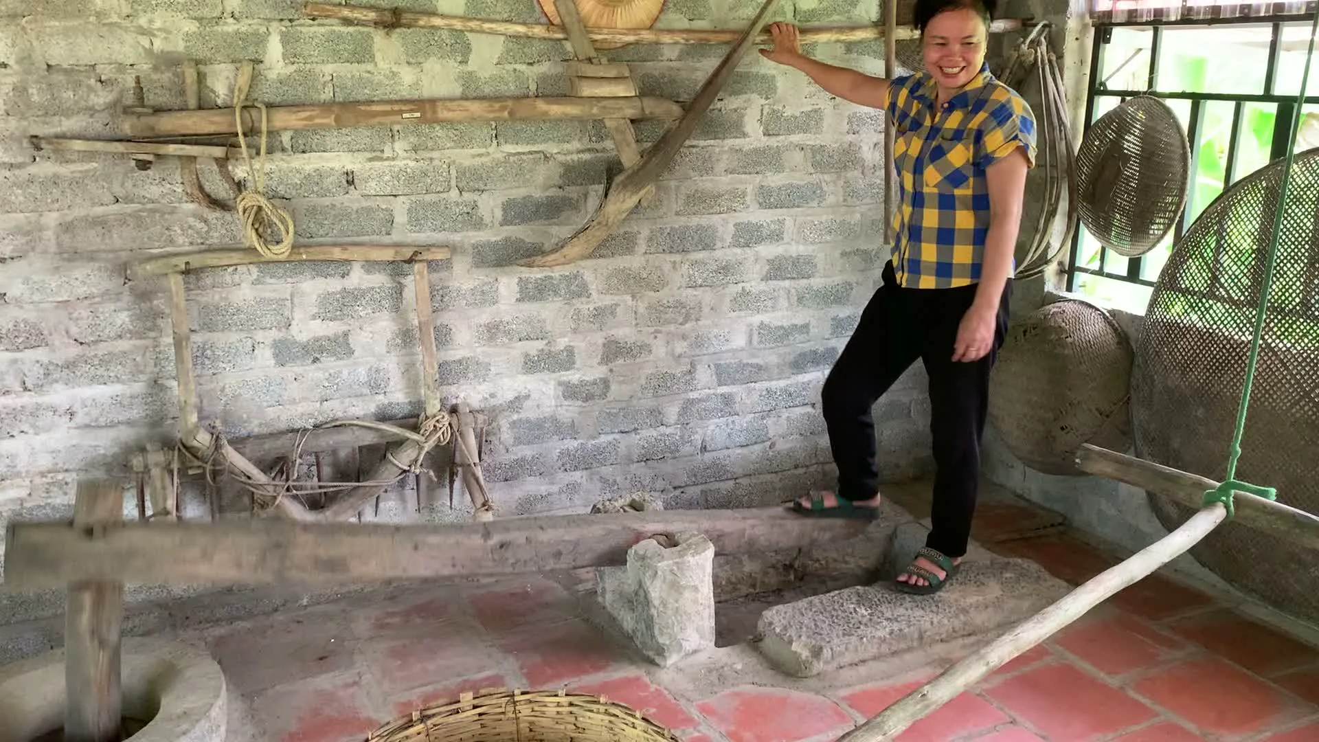 Bối cảnh là bên trong một ngôi nhà hoặc một khu trưng bày, có tường xây bằng gạch xỉ. Một người phụ nữ đang mỉm cười, mặc áo sơ mi kẻ ca rô màu vàng và xanh, quần đen. Cô ấy đang đứng trên một chiếc cối giã gạo bằng gỗ, một nông cụ truyền thống, và dùng chân để vận hành nó. Đây là hoạt động trình diễn cách giã gạo theo kiểu xưa. Xung quanh và treo trên tường có nhiều nông cụ khác như cái bừa, các loại rổ, rá, nia đan bằng tre. Dưới sàn có một chiếc rổ tre.