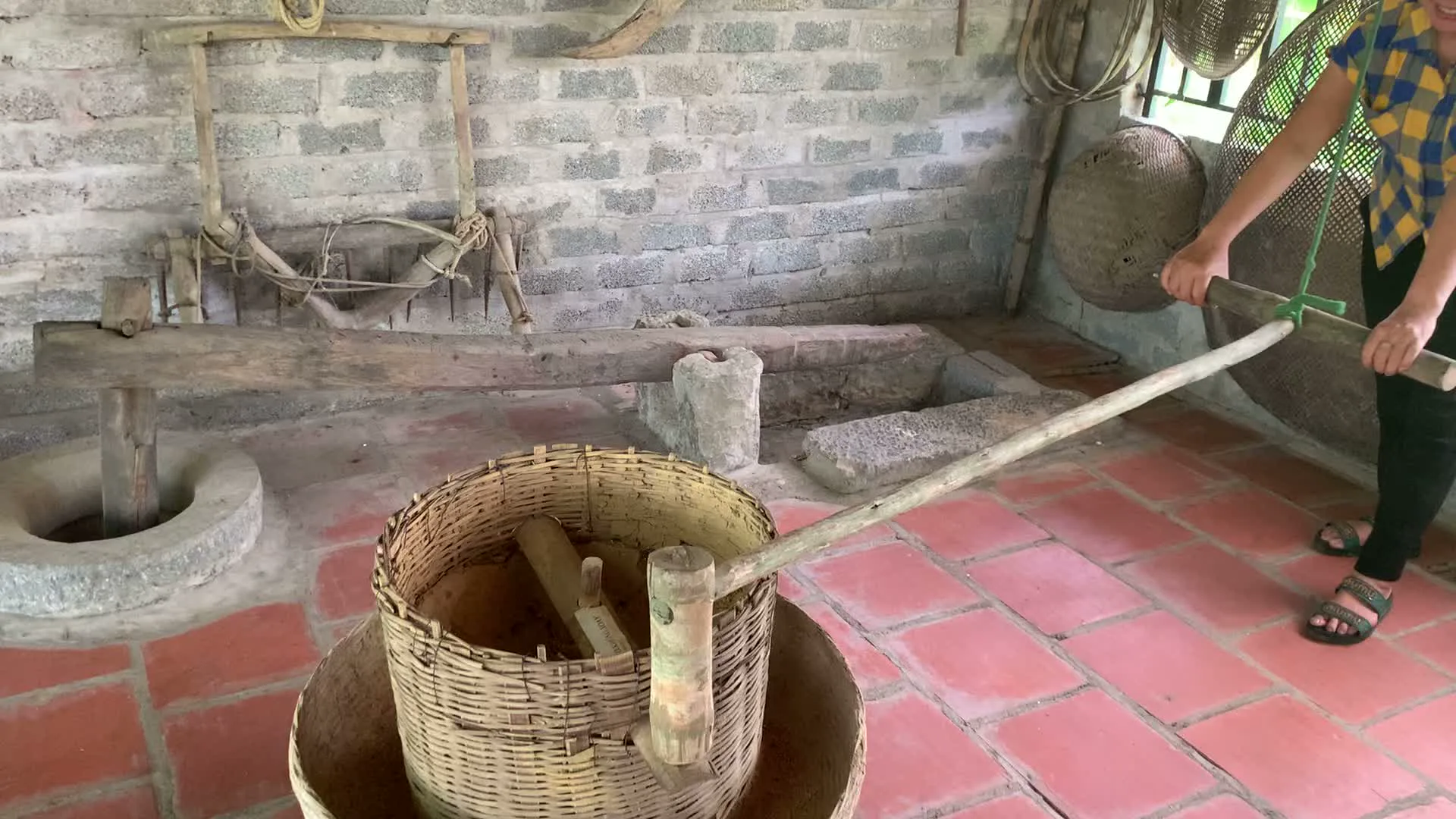 Bối cảnh là một không gian trong nhà, có thể là một ngôi nhà truyền thống hoặc một khu trưng bày, mô phỏng lại đời sống nông thôn xưa ở Việt Nam. Tường nhà được xây bằng gạch xỉ không trát, nền lát gạch đỏ.。Trong một bức ảnh, một người phụ nữ đang thao tác với một chiếc cối xay lúa truyền thống làm bằng đá và gỗ. Bà đang dùng tay đẩy cần gỗ để xoay thớt cối. Phía trên cối có một chiếc rổ tre lớn.。Các bức ảnh khác cho thấy nhiều nông cụ và ngư cụ cổ được treo trên tường như một vật trang trí hoặc trưng bày, bao gồm một chiếc cày bằng gỗ, nón lá, và nhiều loại dụng cụ bắt cá đan bằng tre như nơm, đó. Một chiếc đèn lồng màu đỏ được treo trên trần nhà. Trên các kệ tre, có trưng bày các đồ gốm sứ như chum, vại và bát đĩa. Ngoài ra còn có các bức tranh đóng khung mô tả cảnh sinh hoạt xưa. Một du khách lớn tuổi đang đứng xem các hiện vật.