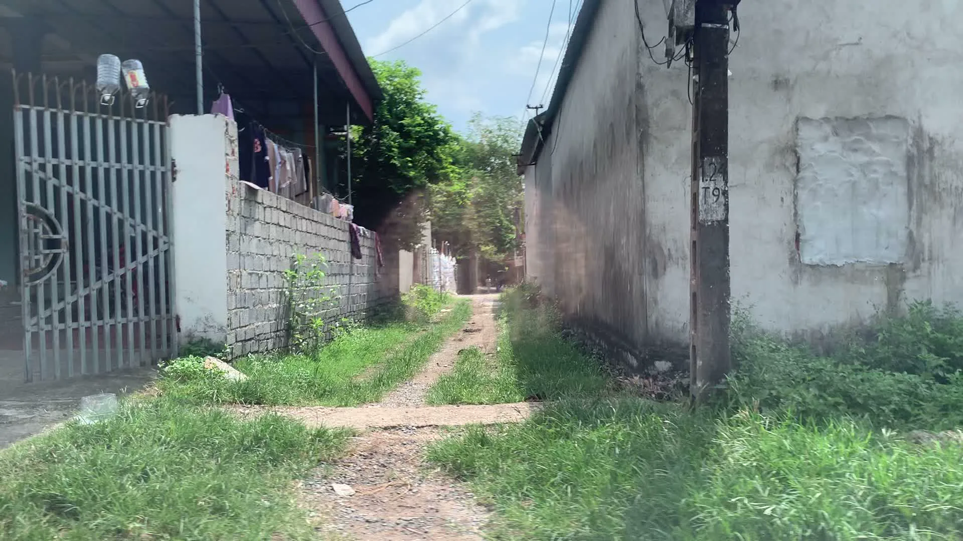 Đây là hình ảnh về một con ngõ nhỏ, đường đất ở một khu dân cư, có thể là ở vùng nông thôn Việt Nam. Con đường mòn, không được lát đá, có cỏ mọc ở giữa và hai bên. Hai bên ngõ là những bức tường nhà. Một bên là ngôi nhà có cổng sắt, tường rào xây bằng gạch, và có mái hiên để phơi quần áo. Bên đối diện là một bức tường bê tông cũ kỹ, trên đó có một cây cột điện với một vài ký tự và số nhưng không rõ ràng, có thể là 'L4 T9'. Một hình ảnh khác cho thấy một bức tường xây bằng gạch block chưa trát, có một cánh cửa sắt nhỏ màu đỏ và một đống cành cây khô chất bên cạnh. Cạnh đó là một cánh cổng sắt lớn hơn cũng màu đỏ, trên đó có vẽ một hình trái tim. Khung cảnh chung mang vẻ yên bình, dân dã của một xóm nhỏ.