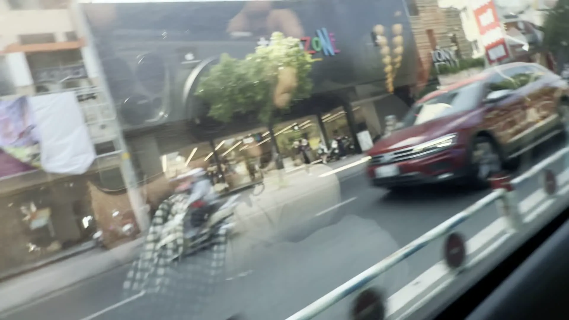 Cảnh quay được ghi lại từ bên trong một chiếc ô tô đang di chuyển trên một con đường trong thành phố. Nhìn qua cửa sổ, có thể thấy một chiếc ô tô SUV màu đỏ đang lưu thông. Ở phía bên kia đường là một tòa nhà hiện đại với mặt tiền bằng kính, trên đó có một tấm biển hiệu lớn ghi chữ "ZONE" với nhiều màu sắc. Một biển hiệu khác bên cạnh có chữ "TONY". Có một vài người và xe máy trên vỉa hè. Ở phía xa bên trái, quần áo được phơi bên ngoài một tòa nhà. Hình ảnh cũng cho thấy cận cảnh nội thất của chiếc xe, bao gồm tay nắm cửa màu bạc và các chi tiết trên tapi cửa.