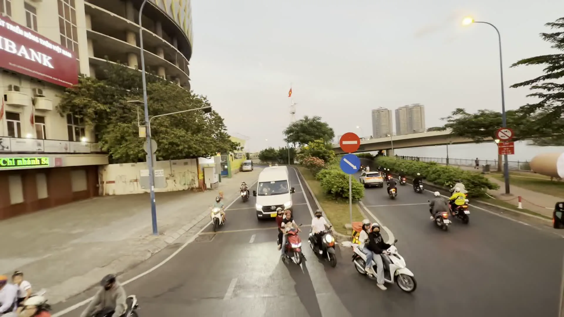 Bản ghi văn bản: Trên tòa nhà bên trái có các dòng chữ "...THÔN VIỆT NAM", "BANK", và "Chi nhánh Sa...". Một tấm biển quảng cáo lớn màu vàng trên hàng rào công trình có nội dung "LIÊN HỆ QUẢNG CÁO" và số điện thoại "0911.39.5555". Các biển báo giao thông có thể nhìn thấy bao gồm biển báo cấm đi ngược chiều, biển báo chỉ dẫn màu xanh và biển báo cấm dừng, đỗ xe.。Mô tả cảnh quan và hoạt động: Đây là khung cảnh giao thông trên một con đường ven sông ở thành phố vào lúc chiều tối. Dòng xe cộ tấp nập với phần lớn là xe máy, ngoài ra còn có ô tô và một chiếc xe van màu trắng. Mọi người đang di chuyển trên đường. Bên trái là một tòa nhà lớn hình tròn đang trong quá trình xây dựng, bên cạnh một tòa nhà khác có vẻ là một ngân hàng. Phía xa có một cây cầu bắc qua sông, và một lá cờ Việt Nam đang tung bay. Đèn đường đã bật sáng. Cây cối được trồng dọc theo lề đường và bờ sông.