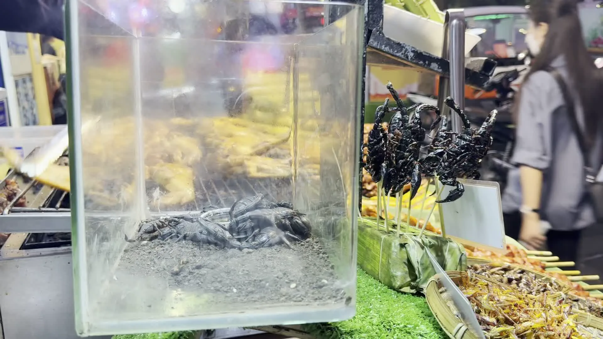 Cảnh quay tại một quầy hàng ăn vặt ở chợ đêm hoặc khu ẩm thực đường phố. Nổi bật ở phía trước là một bể kính, bên trong có nhiều con bọ cạp đen còn sống. Ngay bên cạnh, những xiên bọ cạp đã được chiên giòn được bày bán, cắm trên một khối bọc lá chuối. Ngoài bọ cạp, quầy hàng còn bán nhiều loại côn trùng chiên khác và các món nướng, có thể thấy gà nướng ở phía sau. Khung cảnh cho thấy hoạt động buôn bán tấp nập với người qua lại ở phía sau, trong đó có một phụ nữ đang đeo khẩu trang.