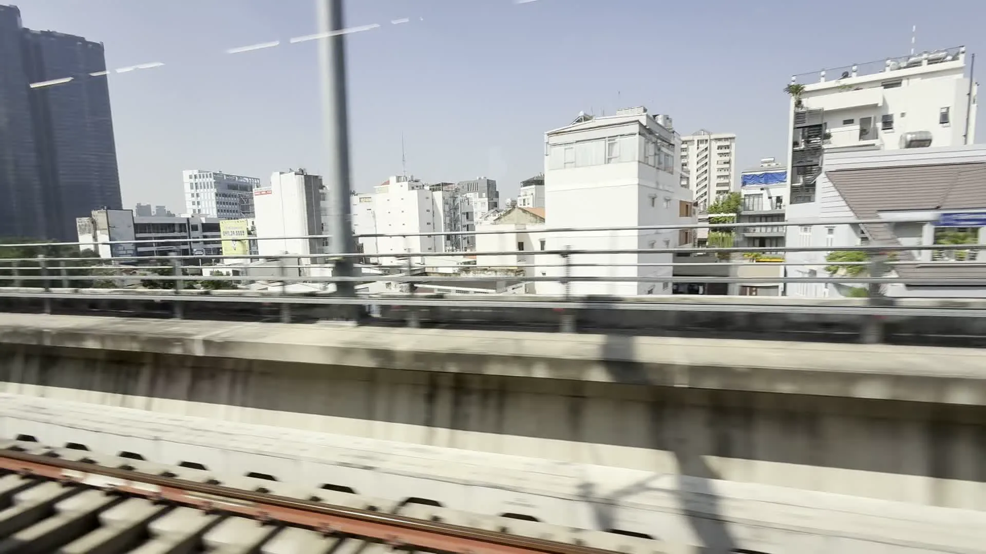 Đây là quang cảnh đô thị được ghi lại từ một đoàn tàu điện trên cao đang di chuyển ở Thành phố Hồ Chí Minh. Ở phía tiền cảnh, có thể thấy đường ray và hàng rào bê tông của tuyến đường sắt. Ở hậu cảnh là một loạt các công trình kiến trúc đa dạng, bao gồm các tòa nhà chọc trời hiện đại, các khu chung cư cao tầng, và những ngôi nhà ở thấp tầng san sát nhau. Nổi bật trong số đó là các tòa nhà cao tầng với mặt ngoài bằng kính và một tòa nhà có kiến trúc hình trụ độc đáo. Xen kẽ giữa các tòa nhà là cây xanh. Bầu trời trong và quang đãng vào ban ngày. Mặc dù có thể thấy một số bảng hiệu trên các tòa nhà ở phía xa, nhưng chữ viết trên đó không thể đọc được do khoảng cách và sự chuyển động của tàu.