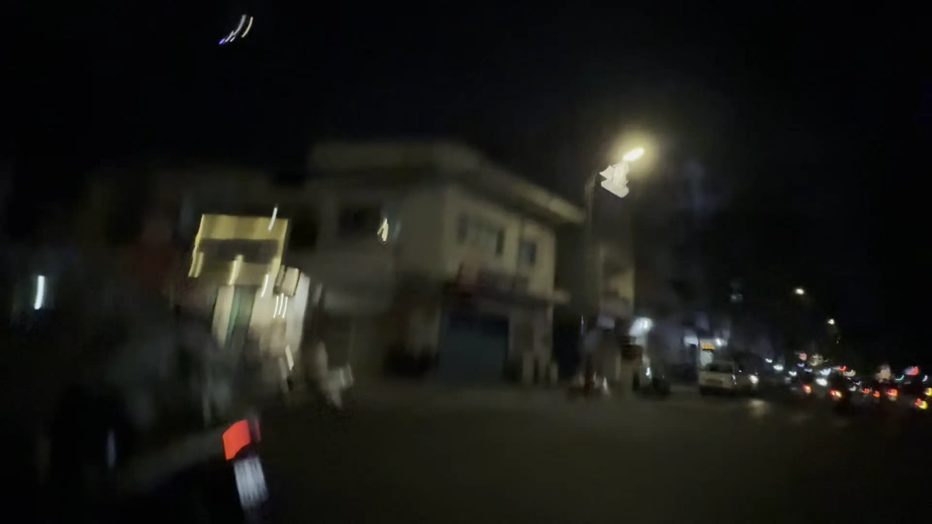 Cảnh quay vào ban đêm trên một con đường trong thành phố, có vẻ như được ghi lại từ một phương tiện đang di chuyển. Hình ảnh cho thấy dòng xe cộ, chủ yếu là xe máy, đang lưu thông trên đường. Nhiều người điều khiển xe máy đội mũ bảo hiểm. Dọc hai bên đường là các tòa nhà nhiều tầng, một số cửa hàng và quán ăn. Có những tòa nhà được trang trí bằng đèn màu rực rỡ. Một cảnh quay khác cho thấy các xe máy đang đi dưới một cây cầu vượt. Có nhiều biển hiệu và một bảng quảng cáo nhưng không thể đọc rõ chữ do hình ảnh bị mờ và thiếu sáng.