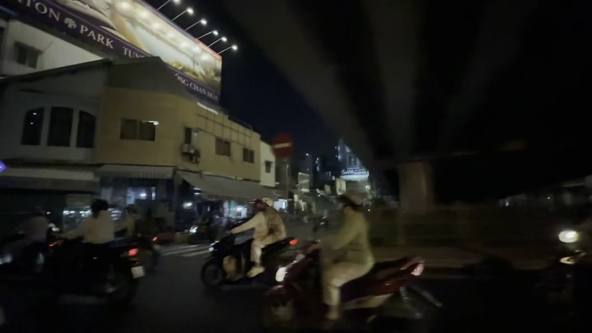 Cảnh quay vào ban đêm trên một con đường đô thị, được ghi lại từ một phương tiện đang di chuyển. Có nhiều người đang điều khiển xe máy trên đường, đội mũ bảo hiểm. Hai bên đường là các tòa nhà cao tầng san sát, đặc trưng của thành phố. Phía trên có một cấu trúc lớn dường như là gầm của một cây cầu vượt. Một biển quảng cáo lớn được chiếu sáng có thể nhìn thấy một phần văn bản, bao gồm các chữ "...TON PARK..." và "...TƯ...". Một biển báo giao thông hình tròn màu đỏ có vạch ngang màu trắng (biển báo cấm đi ngược chiều) cũng xuất hiện trong khung hình. Do ảnh được chụp trong điều kiện thiếu sáng và đang chuyển động, nhiều chi tiết, bao gồm cả chữ viết trên các biển hiệu cửa hàng khác, đều bị mờ và không thể đọc được.