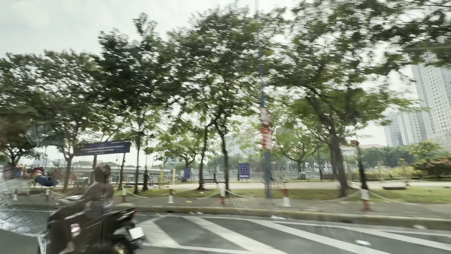 Cảnh quan đường phố được chụp từ một phương tiện đang di chuyển, có thể là ô tô, do đó hình ảnh có phần bị mờ do chuyển động. Khung cảnh cho thấy một con đường nhựa trong đô thị với vạch kẻ đường. Bên lề đường là một khu vực giống như công viên hoặc bờ kè có nhiều cây xanh lớn và ghế đá. Một người đang lái xe máy trên đường. Ở phía xa, có thể nhìn thấy những tòa nhà cao tầng. Có một số biển báo và một cột cờ/biểu ngữ nhưng chữ viết quá mờ để có thể đọc được. Các hình ảnh tiếp theo cho thấy con đường chạy dọc theo một bức tường bê tông cao, trên đỉnh tường có hàng rào và trồng các bụi cây.