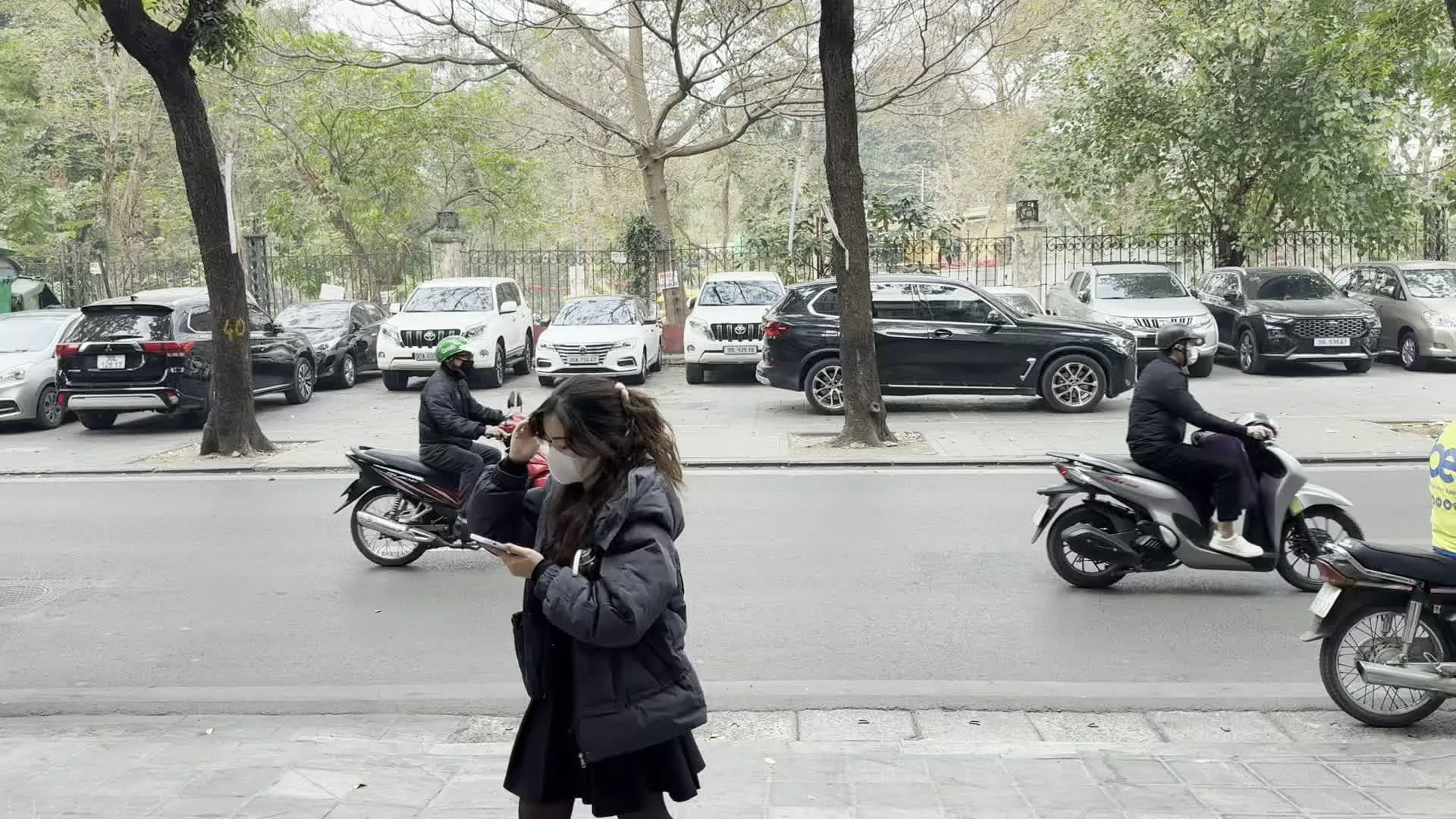 Khung cảnh trên một con phố ở Hà Nội. Bên đường là một bãi đỗ xe với rất nhiều ô tô đang đậu. Dưới lòng đường, nhiều người đang điều khiển xe máy di chuyển. Những người tham gia giao thông đều mặc áo khoác ấm và đội mũ bảo hiểm, cho thấy thời tiết có vẻ lạnh. Một số người có đeo khẩu trang. Trên vỉa hè, một cô gái mặc áo khoác dày đang vừa đi bộ vừa sử dụng điện thoại di động. Cây cối hai bên đường có cành trơ trụi lá. Có một người lái xe máy mặc áo khoác màu vàng, trông giống như một tài xế giao hàng công nghệ.