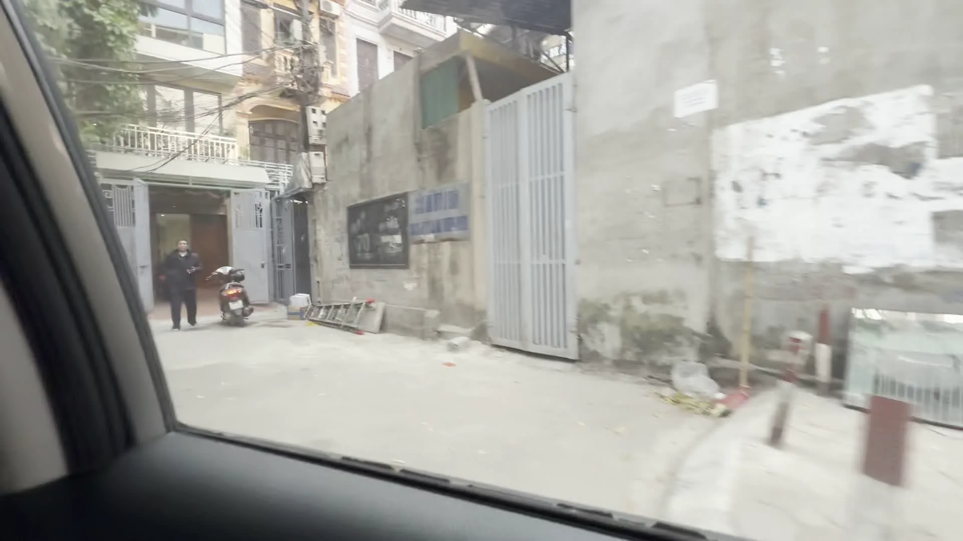 Cảnh quay được ghi lại từ bên trong một chiếc ô tô đang di chuyển trên đường phố Hà Nội. Khung cảnh cho thấy một con ngõ nhỏ đặc trưng của khu dân cư, với những ngôi nhà ống nhiều tầng. Một chiếc xe máy đang đỗ trong ngõ, gần đó có một người đang đứng. Trên bức tường bê tông có gắn một vài tấm biển, nhưng chữ viết trên đó bị mờ, không thể đọc rõ. Khi xe tiếp tục di chuyển, quang cảnh lướt qua một khu vực ven đường được rào lại bằng lưới thép, bên trong chứa nhiều đồ đạc và vật liệu trông như phế liệu hoặc đồ dùng cho công trường. Một tấm áp phích ở khu vực này có thể đọc được một phần chữ 'TÂM AN'. Cuối cùng, hình ảnh lướt qua một cửa hàng có cửa kính, bên trong có sàn lát gạch trắng và một kệ trưng bày, nhưng hình ảnh rất mờ và không thể nhận dạng được chi tiết do xe đang chạy.