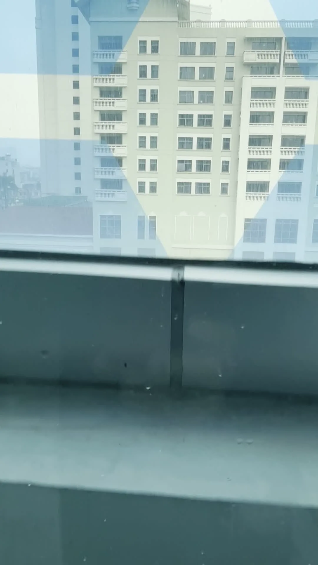 Cảnh quay được ghi lại từ bên trong một tòa nhà, nhìn ra quang cảnh đô thị bên ngoài qua cửa sổ. Ban đầu, hình ảnh cho thấy một tòa nhà cao tầng khác đối diện, có thể là chung cư hoặc khách sạn, với tông màu vàng nhạt và nhiều ban công. Có một hình ảnh phản chiếu mờ trên cửa sổ của một người đeo kính. Sau đó, góc nhìn hướng xuống đường phố, nơi có các phương tiện giao thông như ô tô và một chiếc xe tải phủ bạt đang đỗ. Vỉa hè trông ẩm ướt, cho thấy trời có thể vừa mưa. Xung quanh có cây xanh và các bụi cây được cắt tỉa gọn gàng, tạo nên một khung cảnh đường phố hiện đại ở Hà Nội. Không có chữ viết hay ký hiệu nào có thể đọc được trong các hình ảnh.