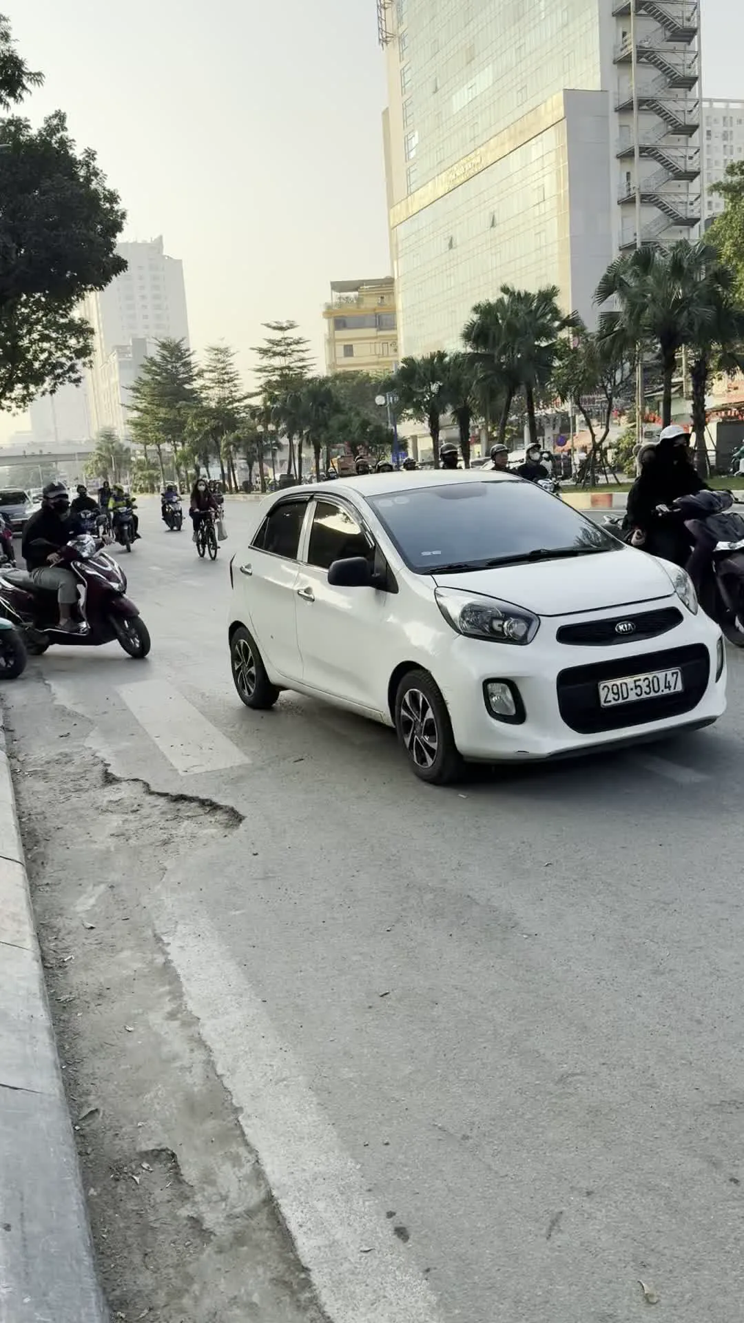 Đây là khung cảnh trên một con đường đô thị ở Hà Nội. Con đường khá rộng, có nhiều làn xe và vạch kẻ đường cho người đi bộ. Hai bên đường là vỉa hè được lát gạch, có cây xanh bao gồm cả cây cọ, và các tòa nhà cao tầng hiện đại. Giao thông trên đường bao gồm cả ô tô và xe máy. Trong ảnh, có một chiếc ô tô Kia màu trắng và một chiếc SUV màu đen đang lưu thông. Nhiều người đang điều khiển xe máy, đội mũ bảo hiểm và đeo khẩu trang. Ở bên lề đường, một người đàn ông đang đứng trên vỉa hè cạnh một chiếc xe máy điện màu xanh. Bề mặt đường nhựa có một số chỗ bị hư hỏng và bong tróc.