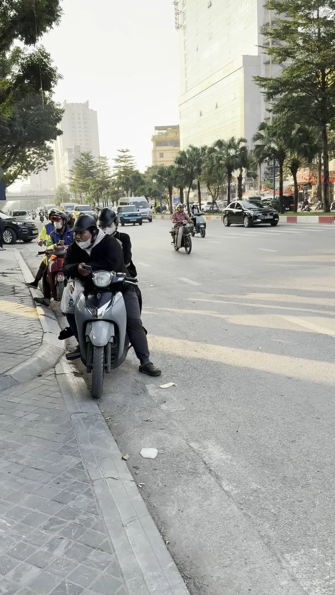 Cảnh đường phố ở Hà Nội với nhiều phương tiện giao thông. Ở bên lề đường, một số người đang dừng xe máy. Một người đàn ông ngồi trên chiếc xe máy màu xám đang sử dụng điện thoại di động. Tất cả những người đi xe máy đều đội mũ bảo hiểm và một số người đeo khẩu trang. Trên đường, dòng xe cộ bao gồm ô tô và xe máy đang di chuyển. Một chiếc ô tô SUV màu đen đang đi qua. Con đường rộng, hai bên có vỉa hè, trồng nhiều cây xanh, và phía sau là các tòa nhà cao tầng. Có các biển hiệu ở hậu cảnh nhưng chữ viết không thể đọc được.