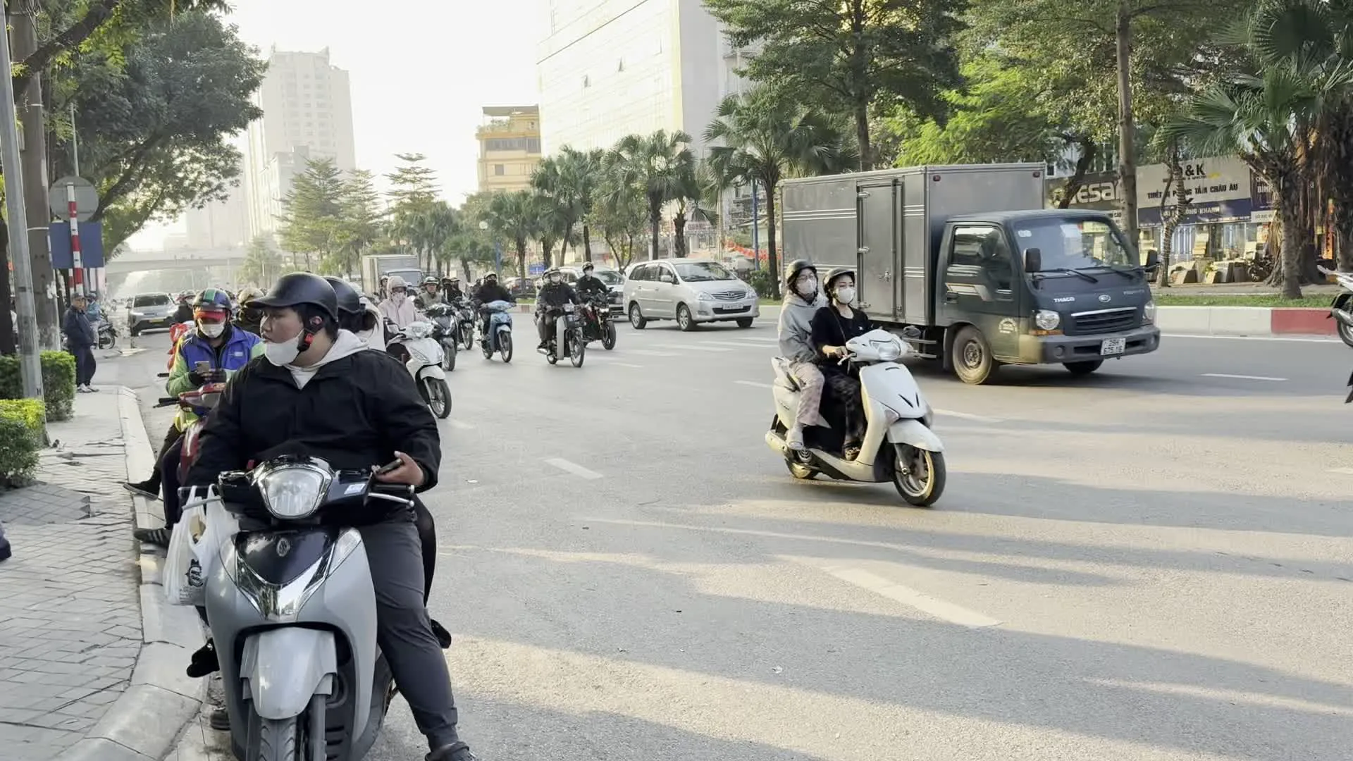 Khung cảnh giao thông đường phố nhộn nhịp vào ban ngày ở một thành phố, có thể là Hà Nội. Rất nhiều người đang di chuyển trên đường bằng phương tiện cá nhân, chủ yếu là xe máy (xe tay ga và xe số). Ngoài ra còn có ô tô, một chiếc xe tải và xe đạp điện. Hầu hết mọi người tham gia giao thông đều đội mũ bảo hiểm, nhiều người đeo khẩu trang. Trong số đó, có một nhóm dường như là học sinh đang đi cùng nhau trên xe máy và xe đạp điện. Hai bên đường là các tòa nhà cao tầng hiện đại và cây xanh, chủ yếu là cây cọ. Có một số biển hiệu quảng cáo nhưng chữ trên đó không rõ ràng, chỉ có thể đọc được một vài từ như 'A&K', 'DESA', 'SỬA CHỮA', 'K&K', 'BIA', 'ĐIỆN THOẠI'.