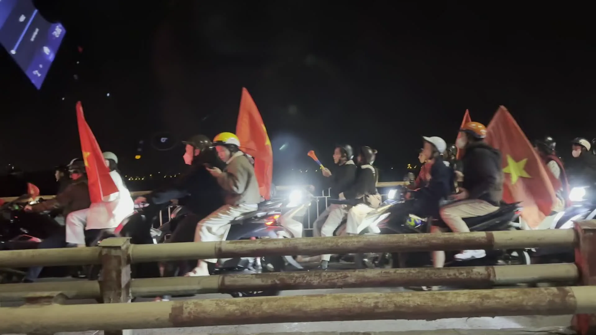 Cảnh tượng diễn ra vào ban đêm trên một cây cầu ở Hà Nội. Một đoàn người đông đúc đang di chuyển bằng xe máy. Nhiều người trong số họ đang cầm và vẫy cờ Tổ quốc Việt Nam (cờ đỏ sao vàng) một cách sôi nổi. Hoạt động này trông giống như một buổi ăn mừng, thường được gọi là "đi bão", có thể là để ăn mừng một chiến thắng thể thao hoặc một sự kiện quốc gia. Hầu hết mọi người đều đội mũ bảo hiểm. Trong một bức ảnh, có người đang cầm một chiếc kèn nhựa. Phía trước là lan can của cây cầu và ở phía xa có thể thấy ánh đèn của thành phố về đêm. Ngoài ra còn có một chiếc ô tô màu trắng di chuyển cùng dòng người. Không có chữ viết nào có thể đọc được trong các hình ảnh.