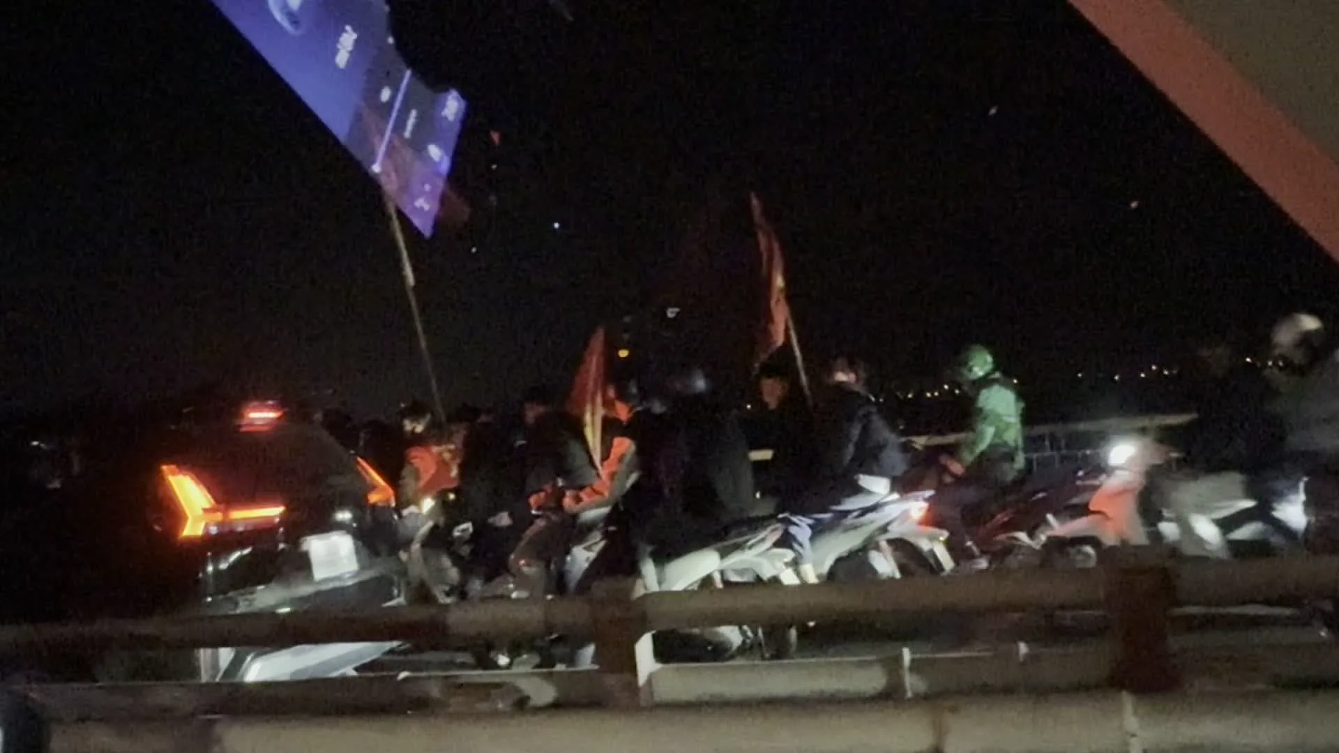 Cảnh tượng vào ban đêm trên một cây cầu ở Hà Nội. Một đám đông người đang đi xe máy và ô tô, nhiều người trong số họ đang cầm và vẫy cờ Việt Nam (cờ đỏ sao vàng). Đây là một hoạt động ăn mừng, thường được gọi là "đi bão", có thể là sau một chiến thắng bóng đá. Mọi người mặc áo khoác và đội mũ bảo hiểm. Dòng xe cộ di chuyển chậm. Ở phía trước có lan can cầu, và phía xa là ánh đèn của thành phố vào ban đêm. Góc trên bên trái của ảnh chụp màn hình giải trí của một chiếc ô tô, cho thấy thời gian là khoảng 22:07.