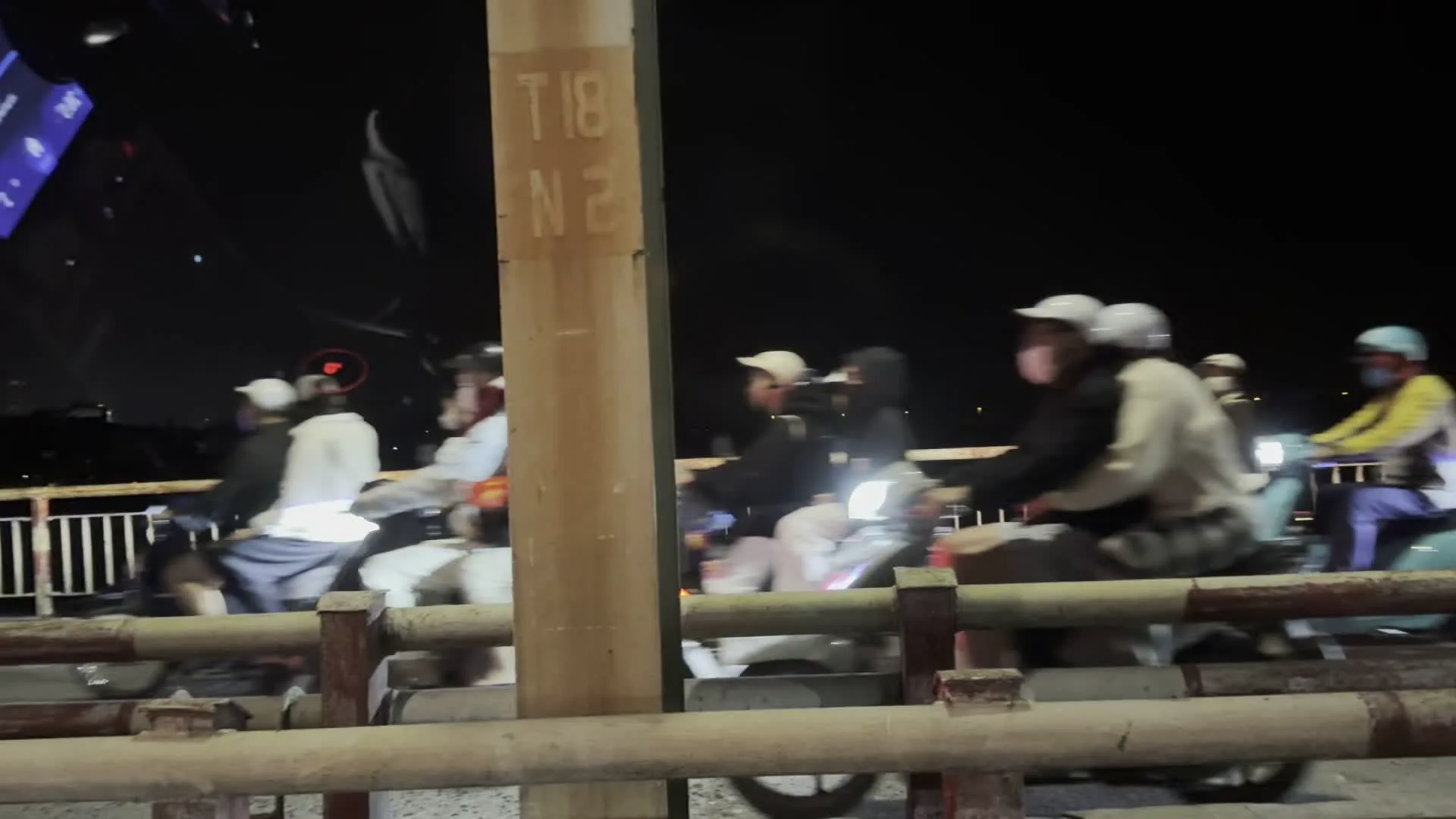 Quang cảnh ban đêm trên một cây cầu ở Hà Nội. Một dòng xe máy rất đông đúc đang di chuyển chậm, có dấu hiệu ùn tắc giao thông. Nhiều người đang điều khiển xe máy trên cầu, hầu hết đều đội mũ bảo hiểm và mặc áo khoác. Phía trước là lan can cầu. Một cột bê tông ở giữa có khắc chữ "T18" và "N2". Đây là một cảnh giao thông thường thấy vào buổi tối tại một thành phố lớn.