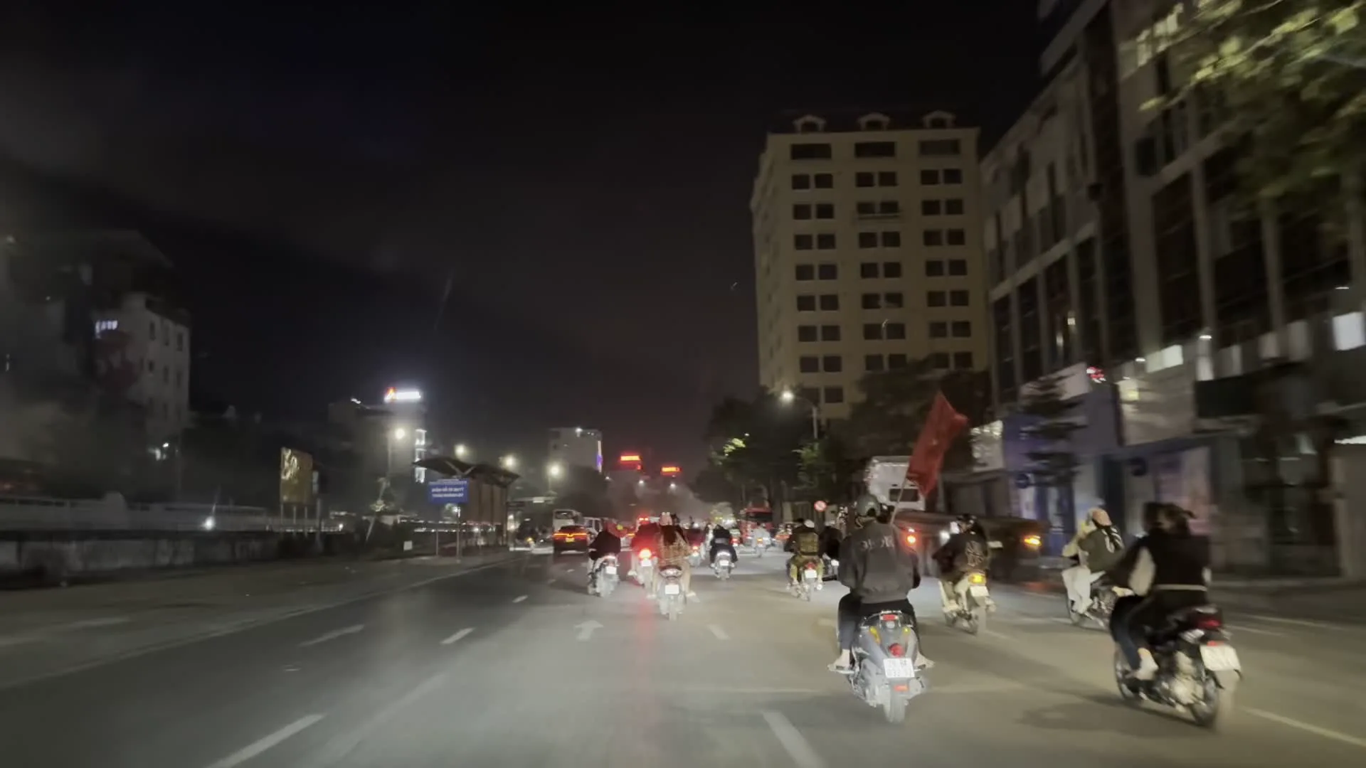Cảnh quay vào ban đêm trên một con đường rộng ở Hà Nội. Một đoàn người đông đúc đang đi xe máy trên đường, trông giống như một cuộc diễu hành ăn mừng. Nhiều người cầm theo cờ đỏ sao vàng của Việt Nam. Hoạt động này thường được gọi là 'đi bão', diễn ra sau một chiến thắng thể thao quan trọng. Hai bên đường là các tòa nhà cao tầng, cây cối và hệ thống đèn đường chiếu sáng. Có thể nhìn thấy một biển hiệu màu đỏ ghi chữ 'HOTEL'. Hầu hết mọi người đều đội mũ bảo hiểm khi tham gia giao thông.