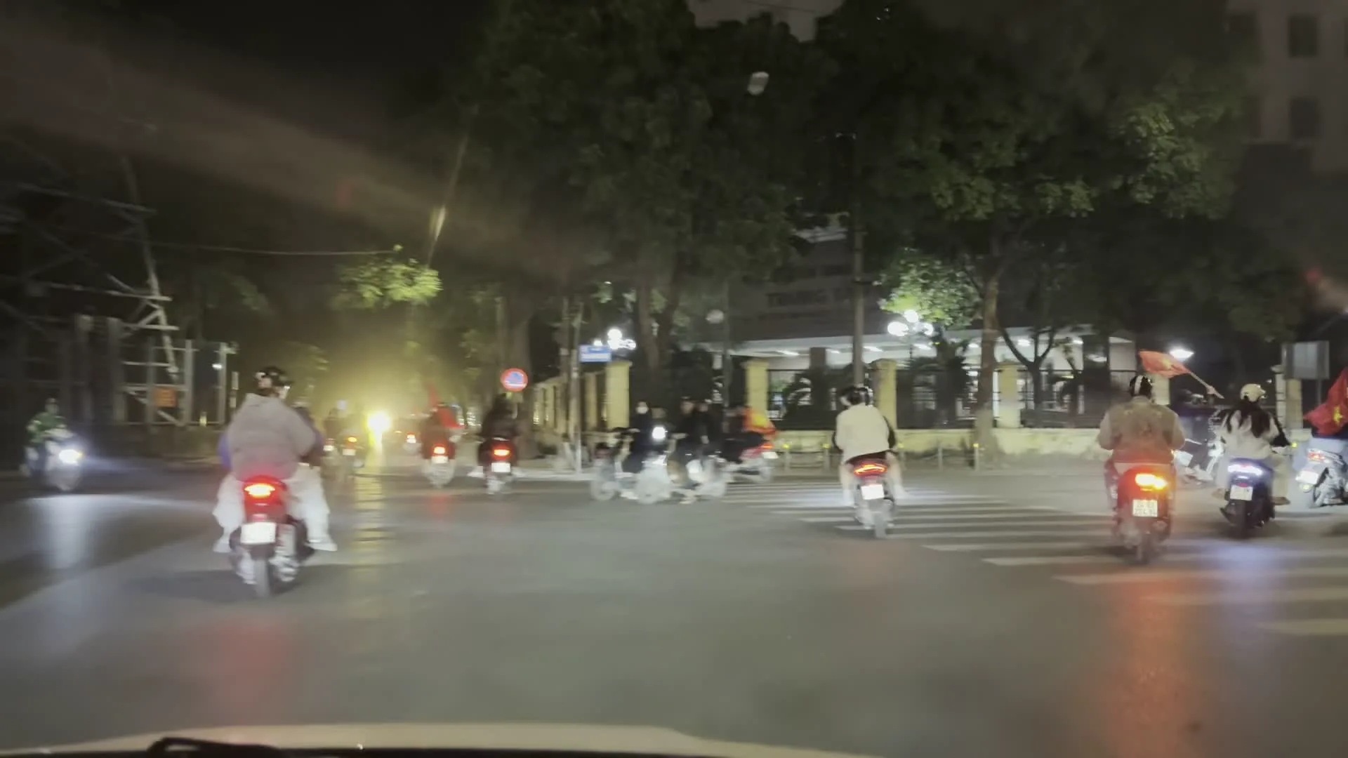Cảnh quay vào ban đêm trên một con đường ở Hà Nội. Rất đông người đang đi xe máy trên đường, tạo thành những đoàn dài. Nhiều người trong số họ, bao gồm cả người lái và người ngồi sau, đang cầm cờ đỏ sao vàng của Việt Nam. Hoạt động này cho thấy một không khí ăn mừng công cộng, thường được gọi là "đi bão" ở Việt Nam, để ăn mừng một sự kiện quan trọng, chẳng hạn như một chiến thắng thể thao. Con đường được chiếu sáng bởi đèn đường và hai bên là hàng cây và các tòa nhà. Trong một số khung hình, có thể thấy các biển hiệu trên tòa nhà và biển báo giao thông, nhưng chữ viết và biểu tượng không thể nhận dạng rõ ràng do điều kiện ánh sáng và chuyển động.