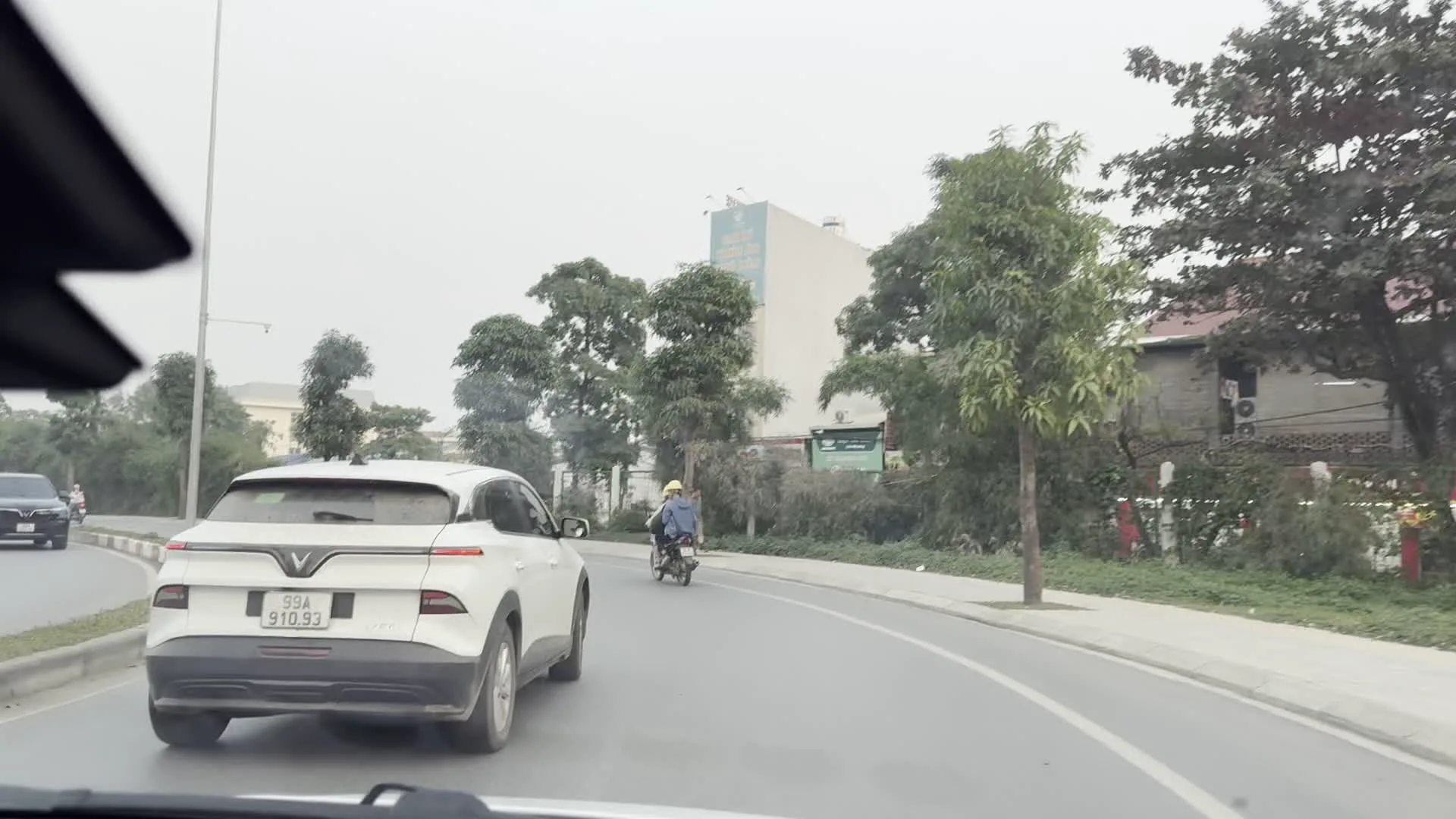 Hình ảnh ghi lại khung cảnh giao thông trên các con đường ở khu vực Hà Nội, được quan sát từ bên trong một chiếc xe hơi đang di chuyển. Các bức ảnh cho thấy sự đa dạng của phương tiện giao thông, bao gồm một chiếc ô tô SUV màu trắng của hãng VinFast, nhiều xe máy, và một chiếc xe tải hạng nặng đang vận chuyển một cỗ máy xây dựng lớn màu xanh lá. Một người đi xe máy mặc áo khoác có dòng chữ 'BUBBLE BUBBLE'. Phong cảnh hai bên đường thay đổi, từ những con đường có cây xanh và các tòa nhà dân cư, đến các đại lộ hiện đại với hàng cây cọ được trồng ở dải phân cách. Một tòa nhà lớn ven đường có biển quảng cáo với dòng chữ 'KIMS'. Bầu trời trong các bức ảnh đều u ám và có nhiều mây.